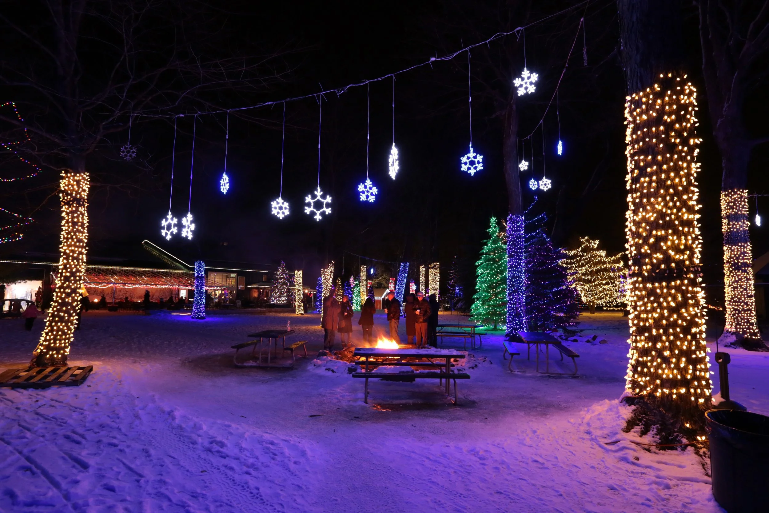 Nighttime scene of a winter outdoor Christmas celebration with decorated trees wrapped in lights, hanging snowflake ornaments, and a group of people gathered around a fire pit in snow-covered ground.