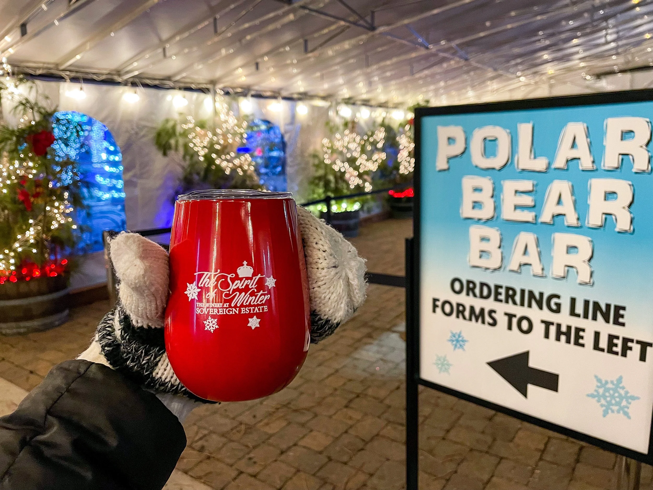 Hand holding a red festive mug with the text "The Spirit of Winter, The Winery at Sovereign Estate." in front of a Christmas market with decorated trees and a lit Christmas ambiance. A sign indicates the Polar Bear Bar with an arrow pointing left, an