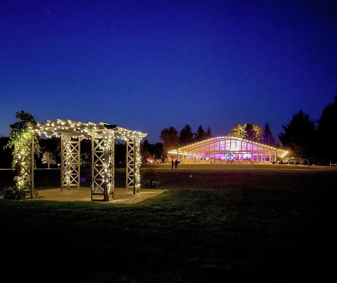 Outdoor evening scene with a wooden pergola adorned with string lights and an illuminated modern building in the background, set against a deep blue sky.
