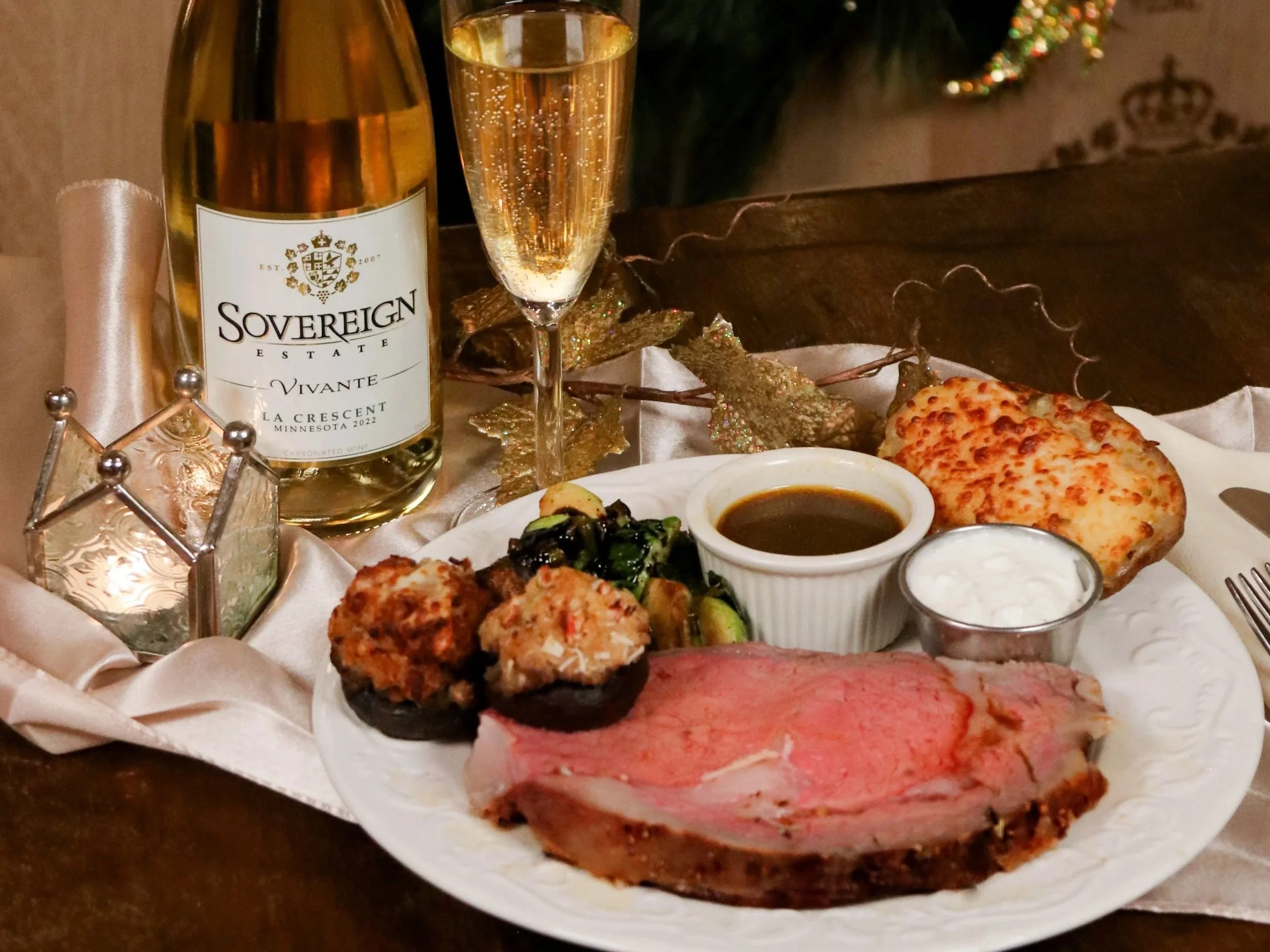 A table setting with a bottle of Sovereign wine, a glass of champagne, and a plate of various foods including bread, meats, and salads, with holiday decorations in the background.