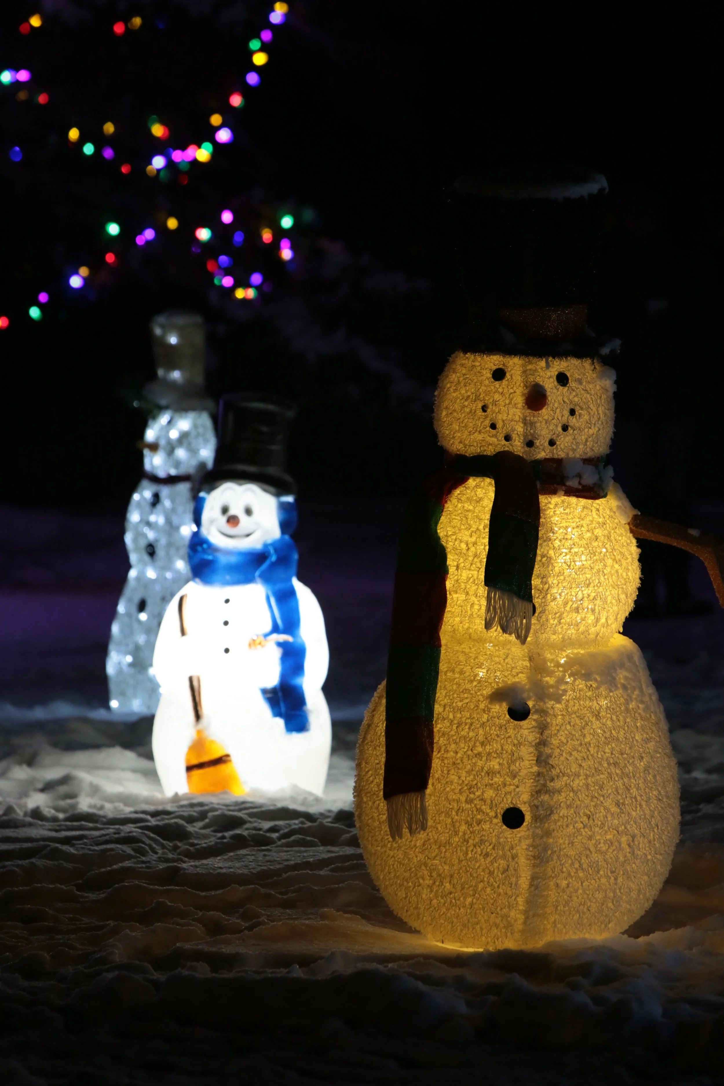 Illuminated snowmen decorations at night, one with a black top hat and a scarf, set against blurred Christmas tree lights.