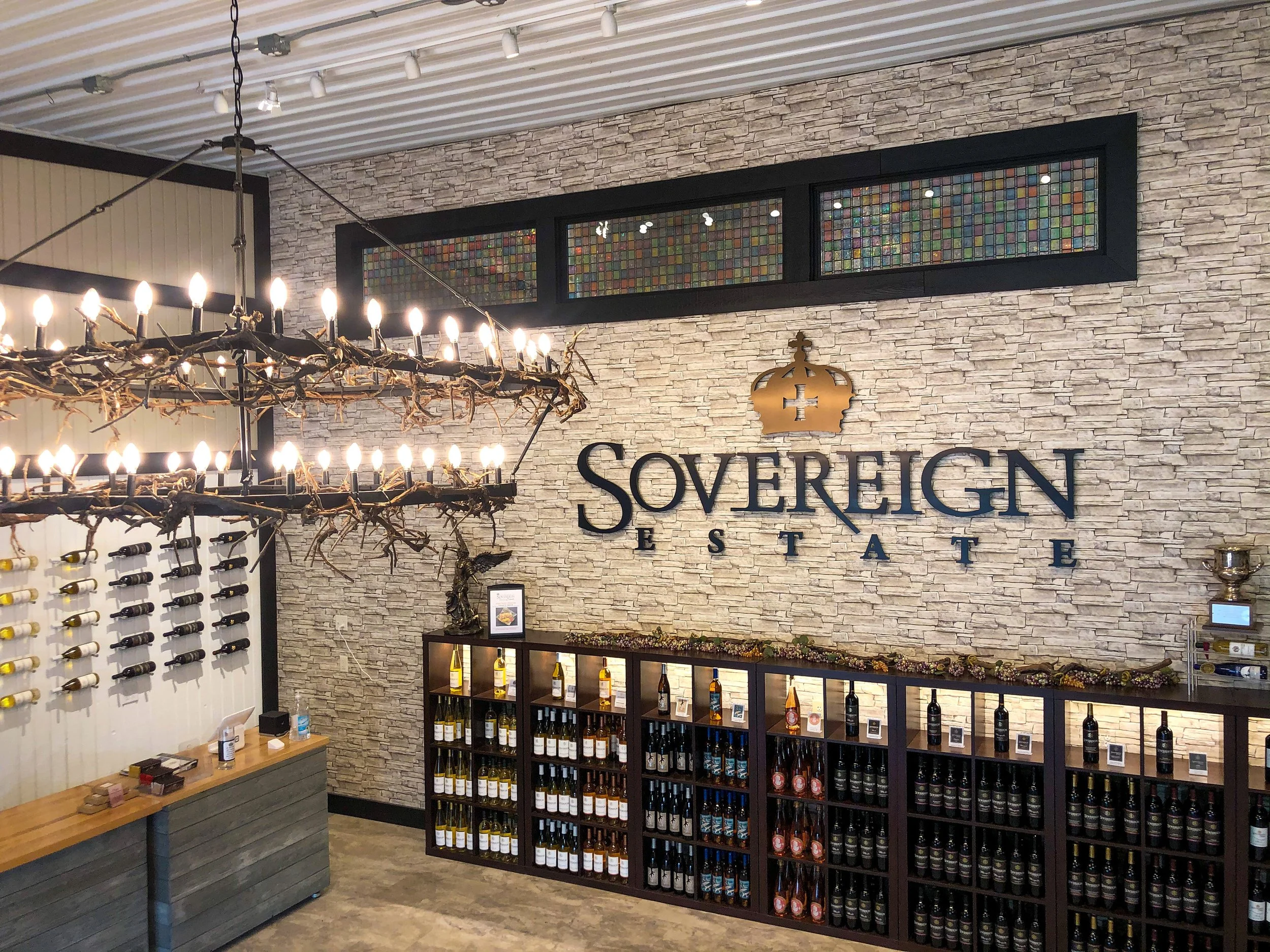 Interior of Sovereign State wine store with wine bottles on shelves, a chandelier made of twigs and candles, and a decorative brick wall with a gold crown logo.