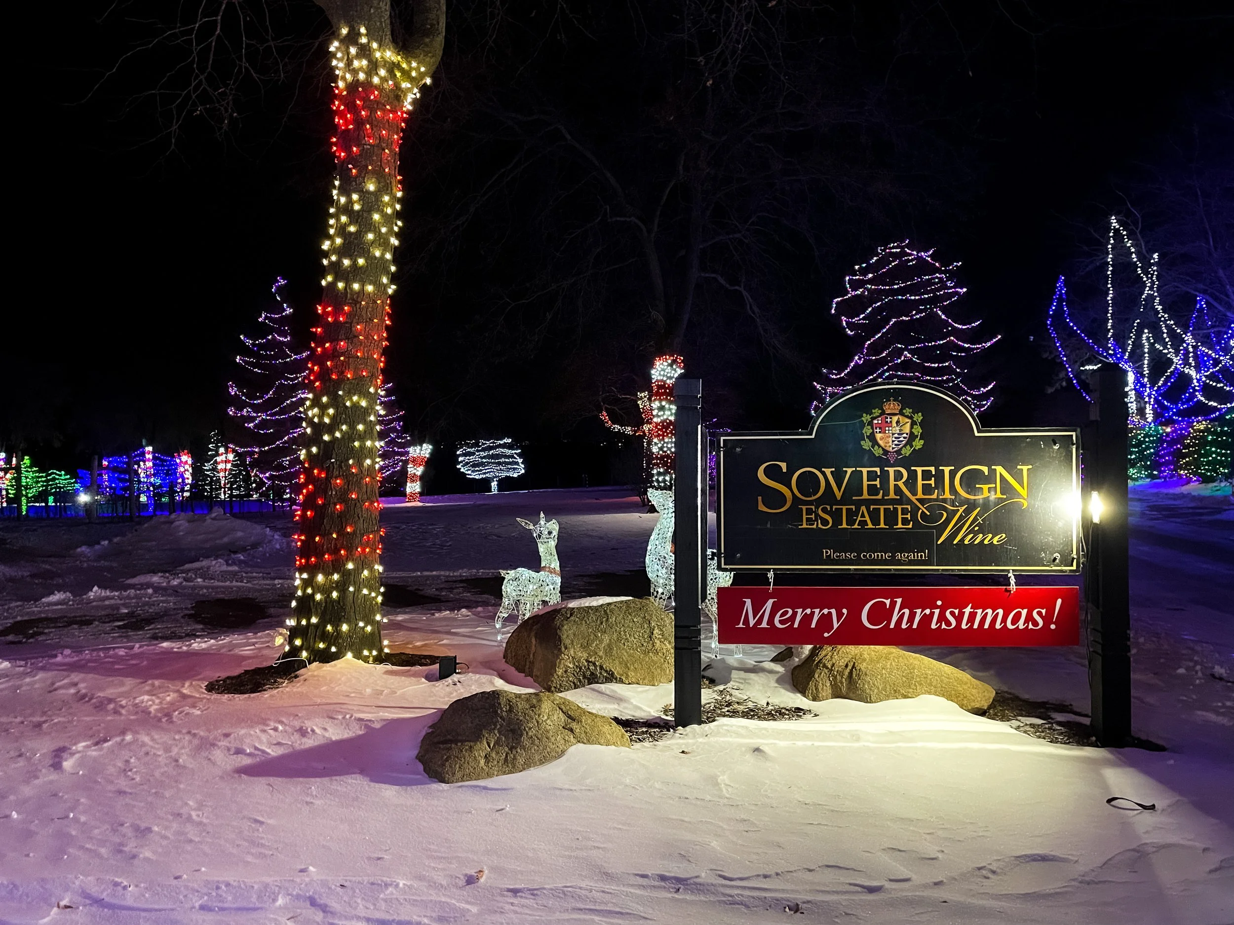 Decorated Christmas trees with colorful lights, illuminated reindeer figures, and a sign that reads 'Sovereign Estate Wine, Please come again! Merry Christmas!' set in a snowy landscape at night.