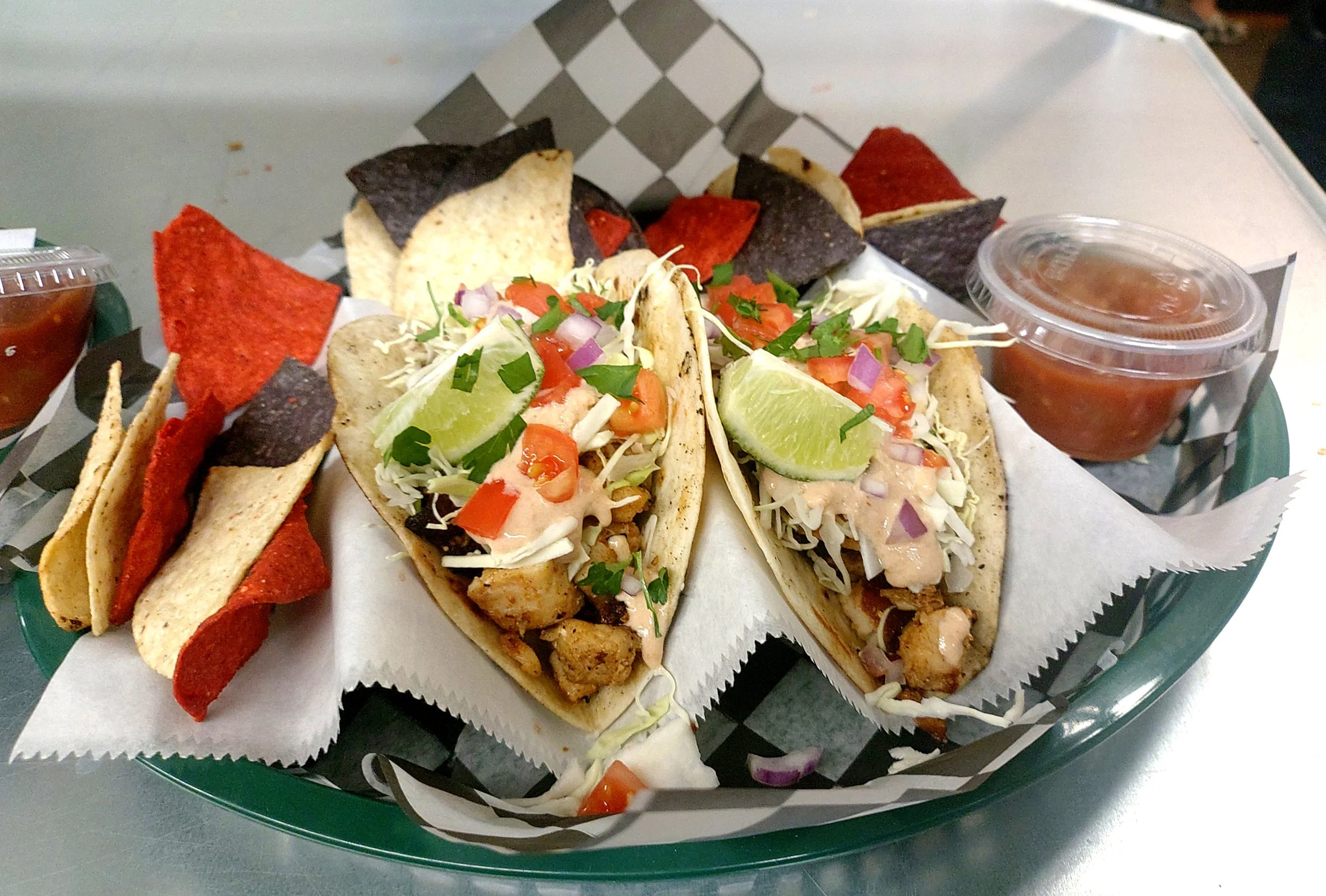 Two chicken tacos topped with lettuce, tomatoes, onions, cilantro, and lime wedges on a basket lined with checkered paper, accompanied by assorted tortilla chips and salsa in plastic containers.
