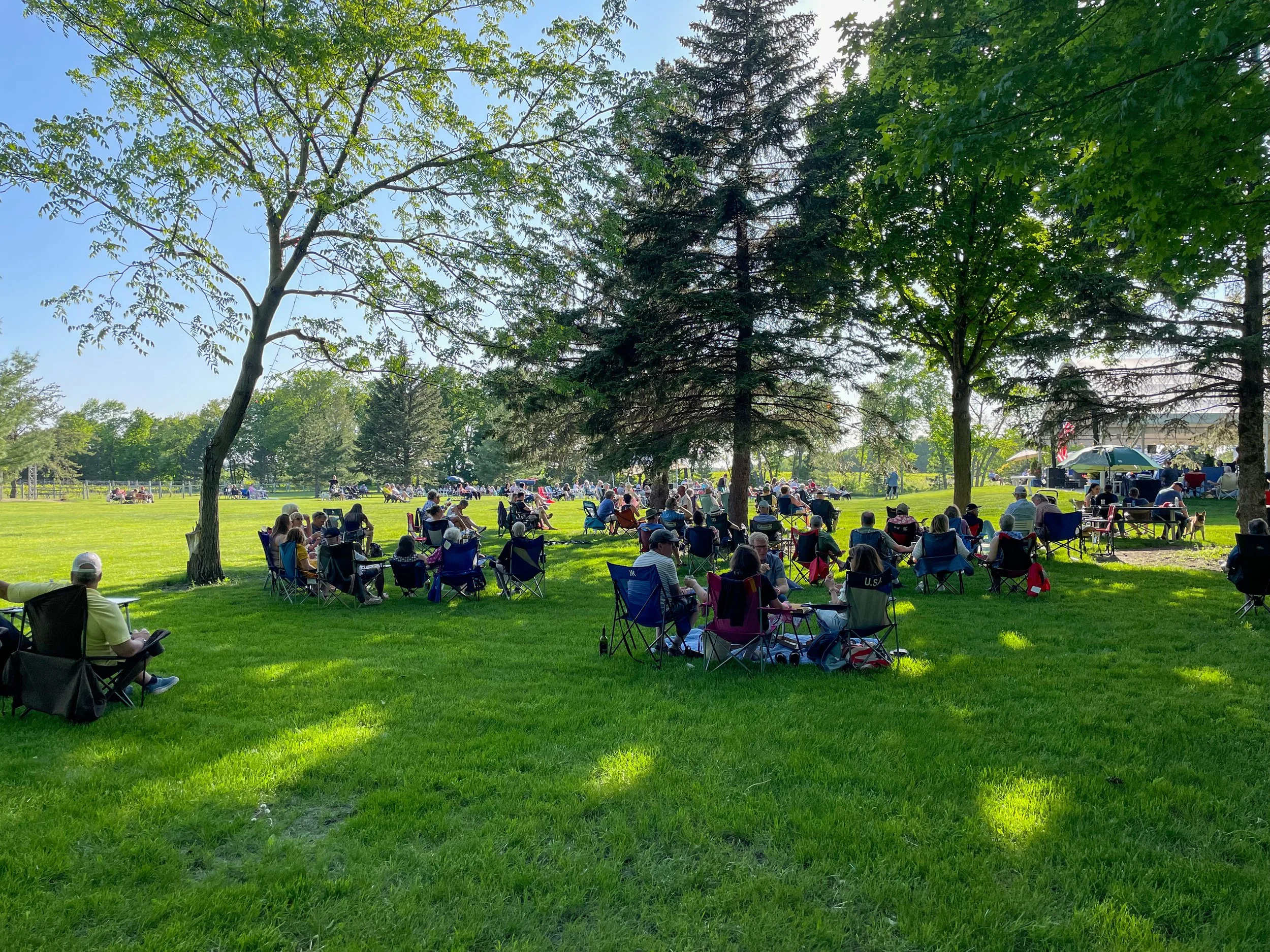 People gathered outdoors on a grassy area sitting in chairs, listening to a live performance near trees.