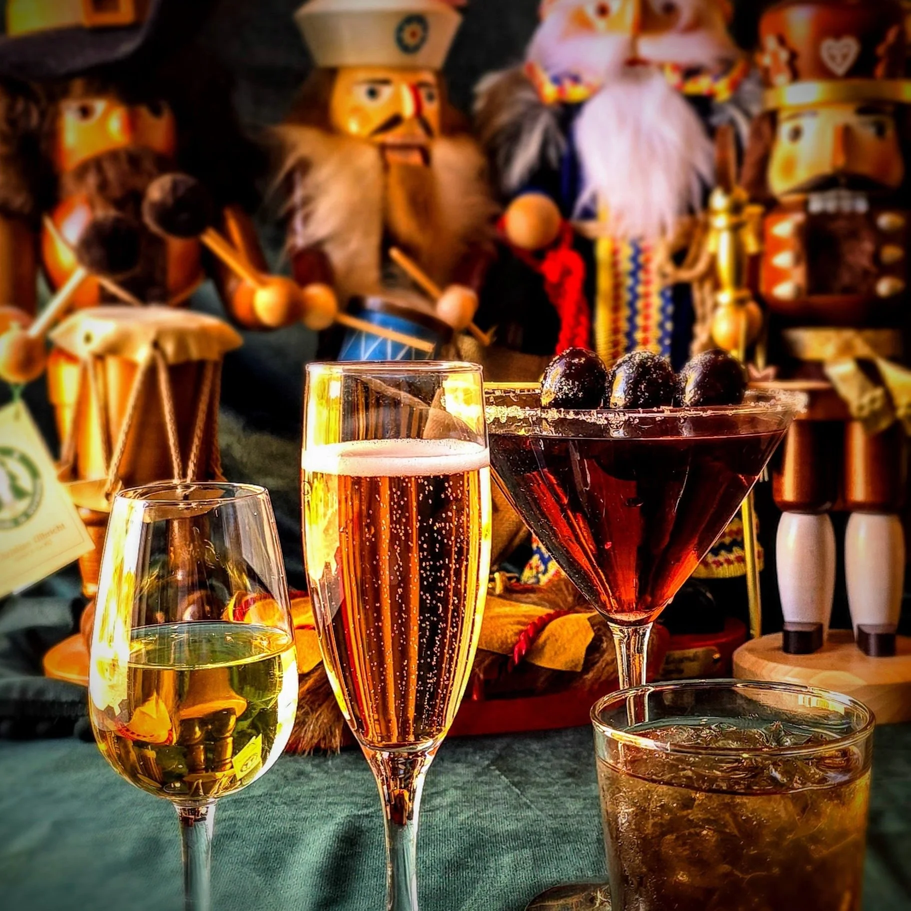 Four drinks on a table in front of traditional Norwegian troll and folk art decorations, including a glass of white wine, a glass of sparkling wine, a cocktail with cherries, and a soda with ice.