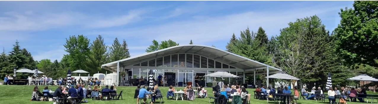 A large outdoor gathering at a park with a modern pavilion, people sitting at tables, enjoying food and drinks, with trees and blue sky in the background.