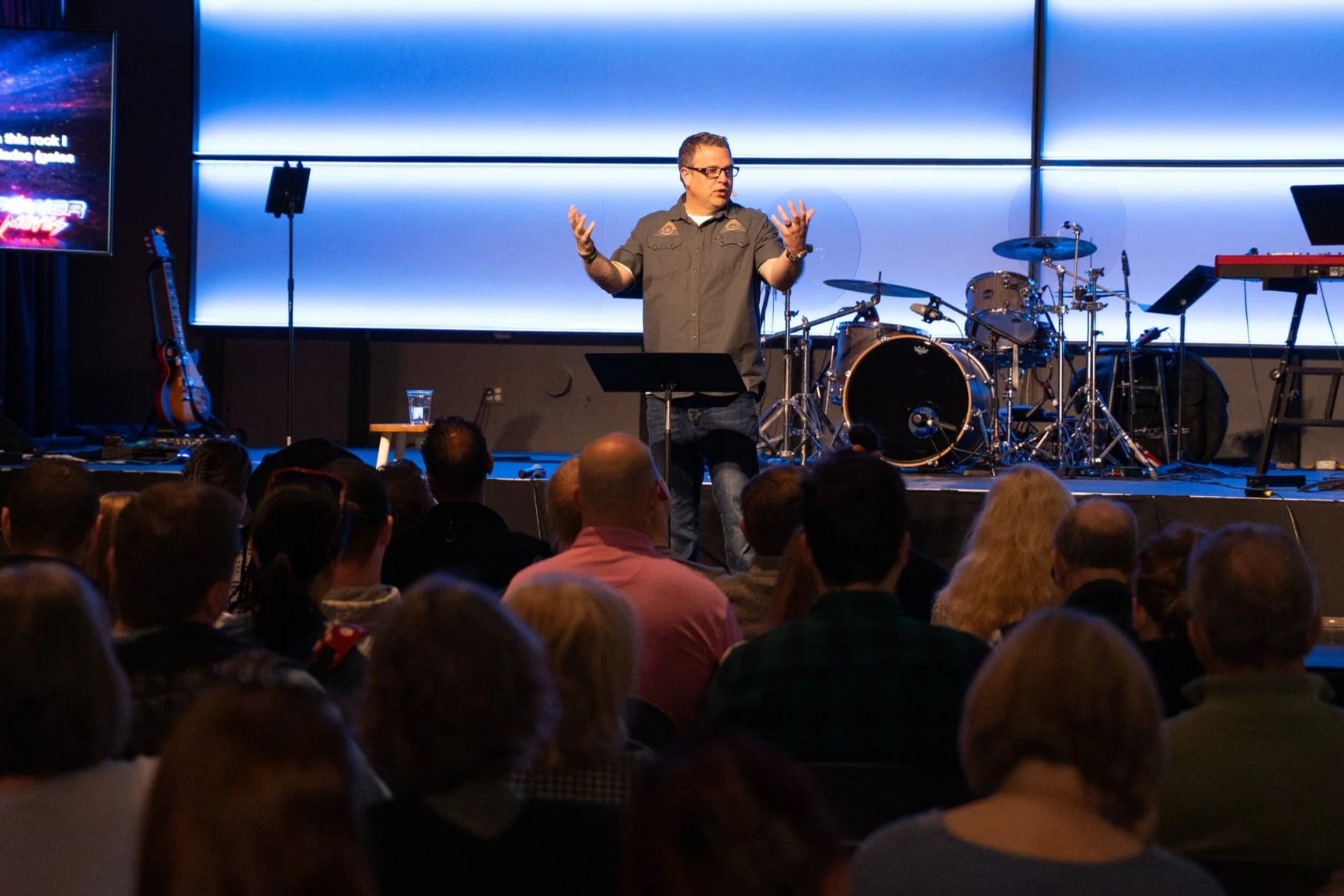 A speaker on stage holding a microphone with a band in the background, next to a wooden cross and a screen displaying text. The setting appears to be a church or worship venue.