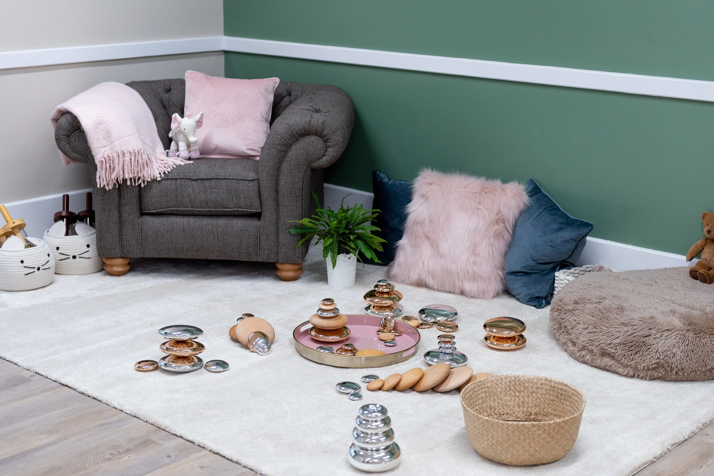 Children's play area with a beige rug, wooden stacking toys, a pink tray with glass stones, plush pillows, a stuffed rabbit, a plant on a white pot, and a brown pet bed with a teddy bear, against a green and cream wall.