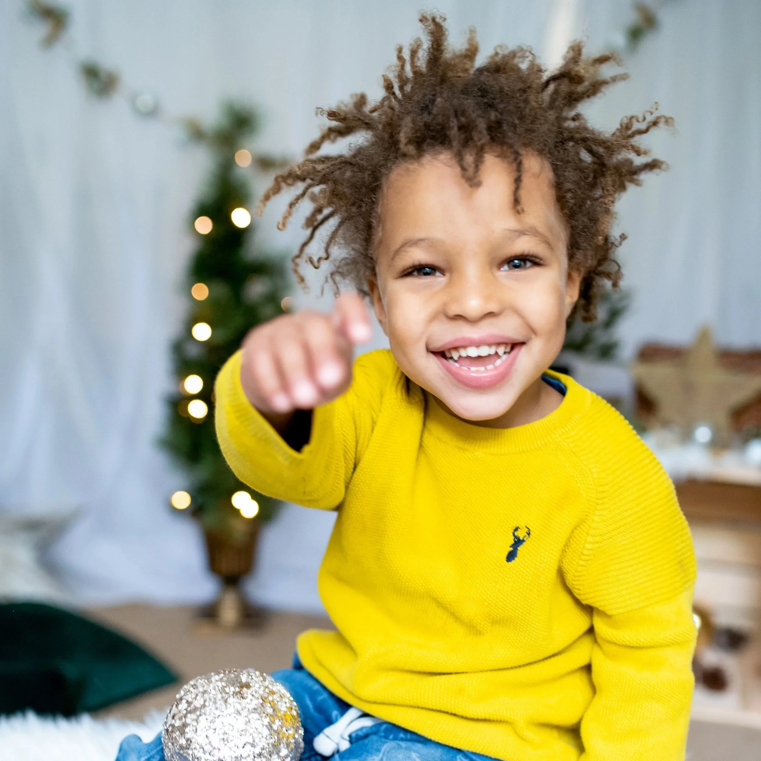 A young boy with curly hair, wearing a yellow sweater, smiling, and reaching towards the camera. He is sitting on a fluffy white surface surrounded by holiday ornaments, with a decorated Christmas tree in the background.