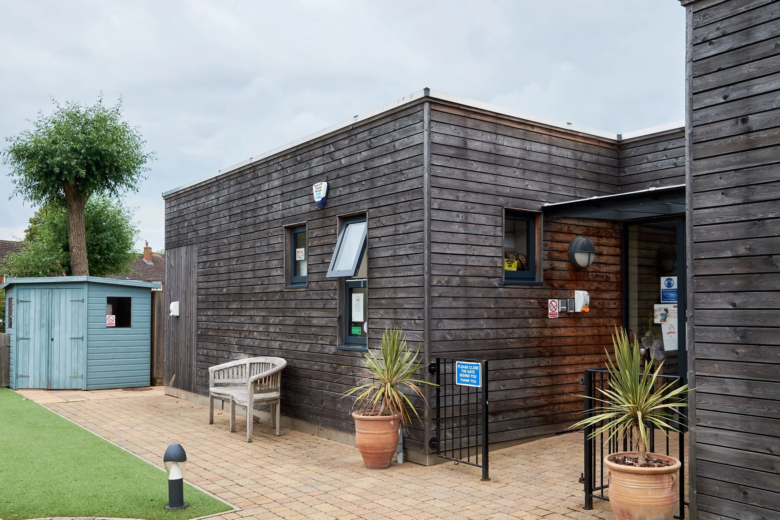 A small dark wooden building with a window, door, and security cameras, next to a blue shed with a sign that says no smoking, surrounded by potted plants and a bench, on a paved walkway with a patch of artificial grass.