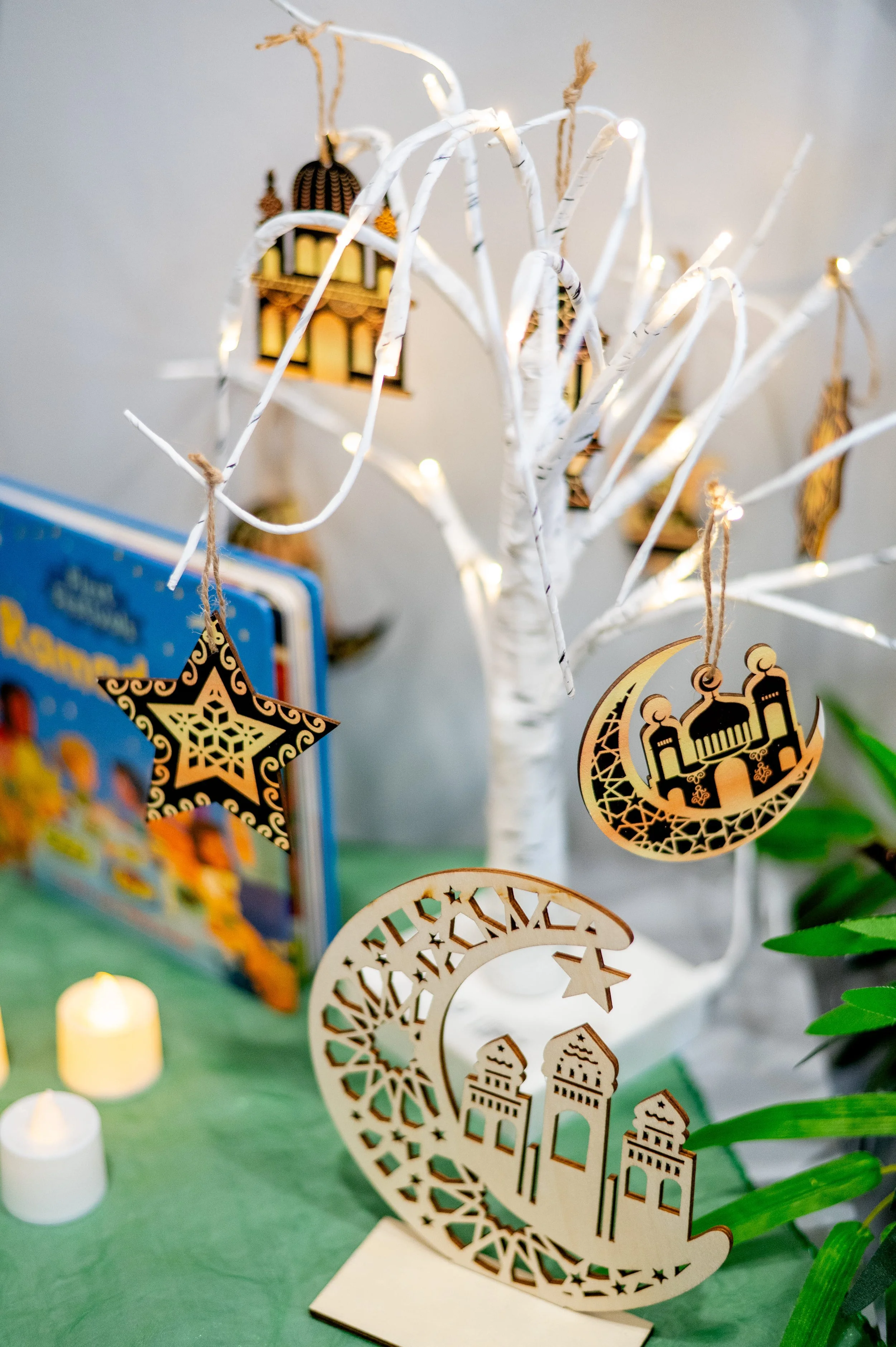 Decorative ornaments hanging from a white, leafless tree, with Islamic motifs, including a mosque and crescent moon, and in the background a partially visible book and lit candles.