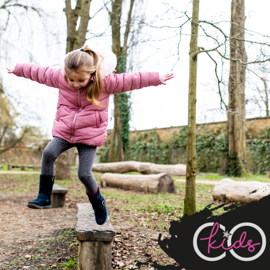 A young girl in a pink coat balancing on a wooden beam in a park.