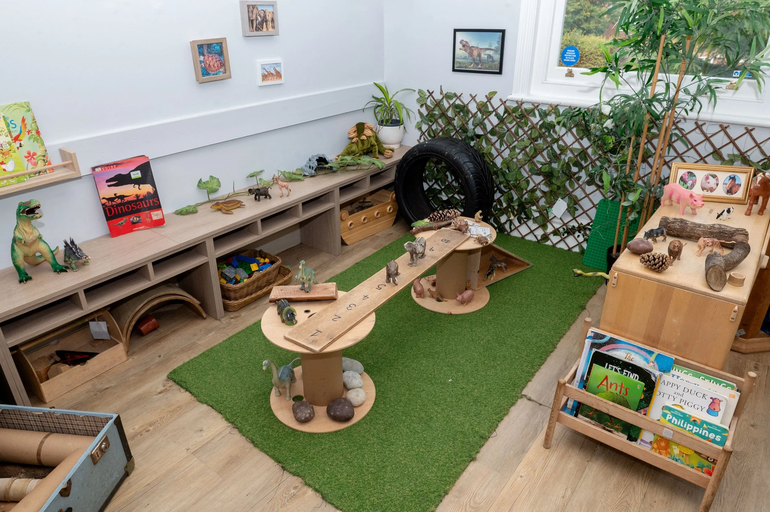 Children's playroom with toy animals, books, and play furniture, featuring a green carpet, a wooden shelves with animal toys, illustrated books, and decorations, and a window with plants.