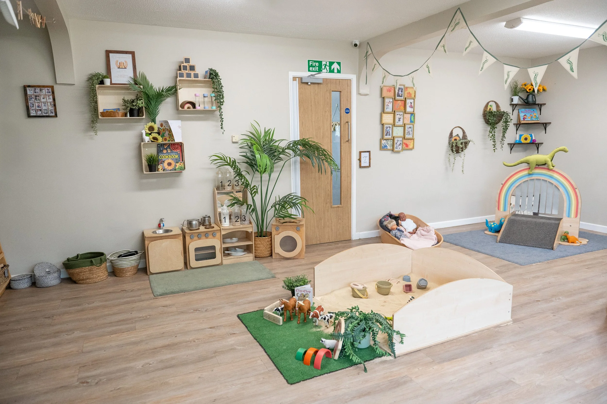 A children's playroom with wooden toys, stuffed animals, toys on a green mat, shelves with books and plants, and a small play kitchen area. There's a colorful rainbow arch with a dinosaur stuffed toy on top, and framed pictures on the wall.