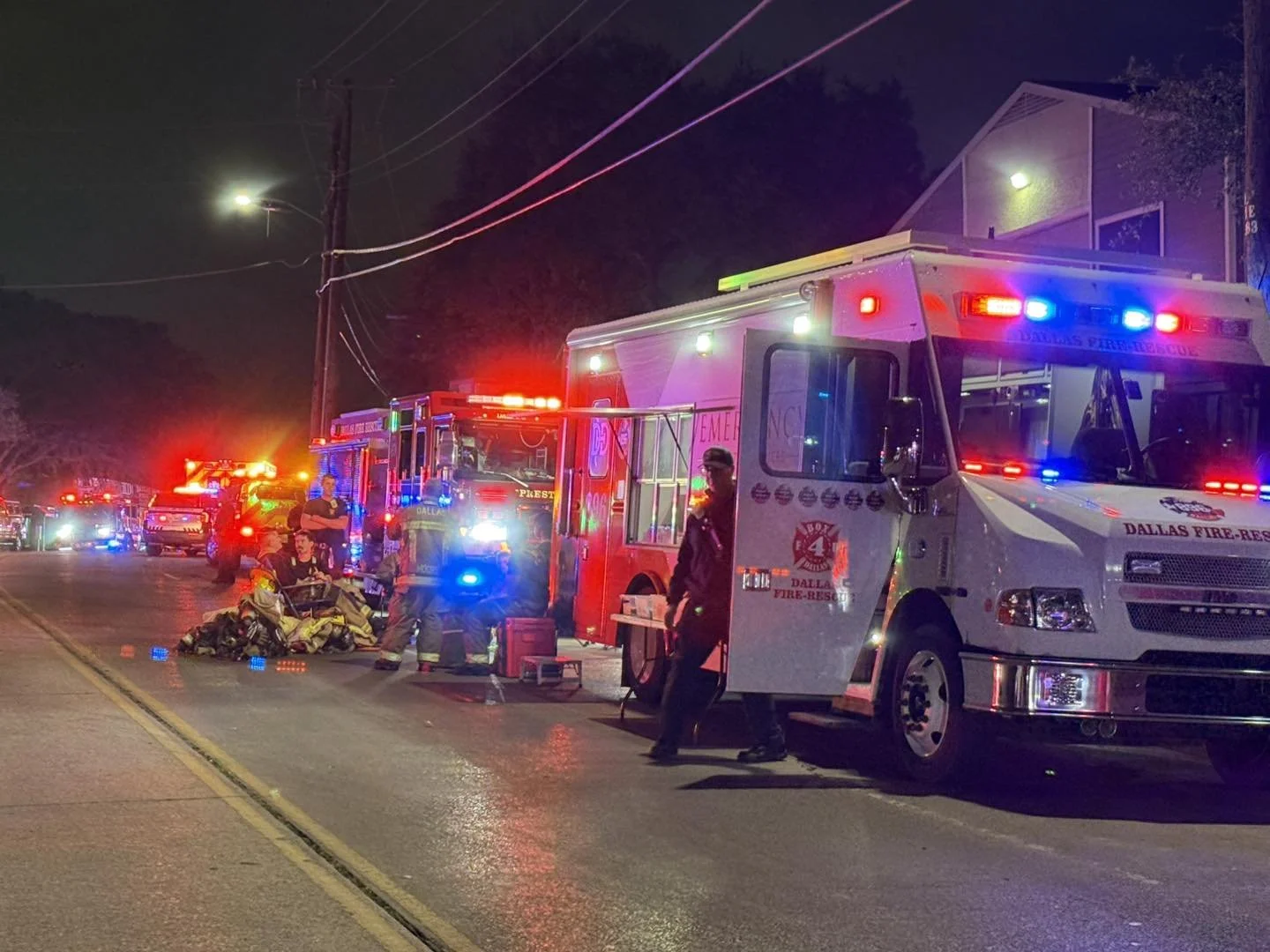 Nighttime scene with emergency vehicles from Dallas Fire-Rescue, including fire trucks and ambulances, with flashing red and blue lights on a residential street. Firefighters are attending to a person on a stretcher, and other emergency personnel are