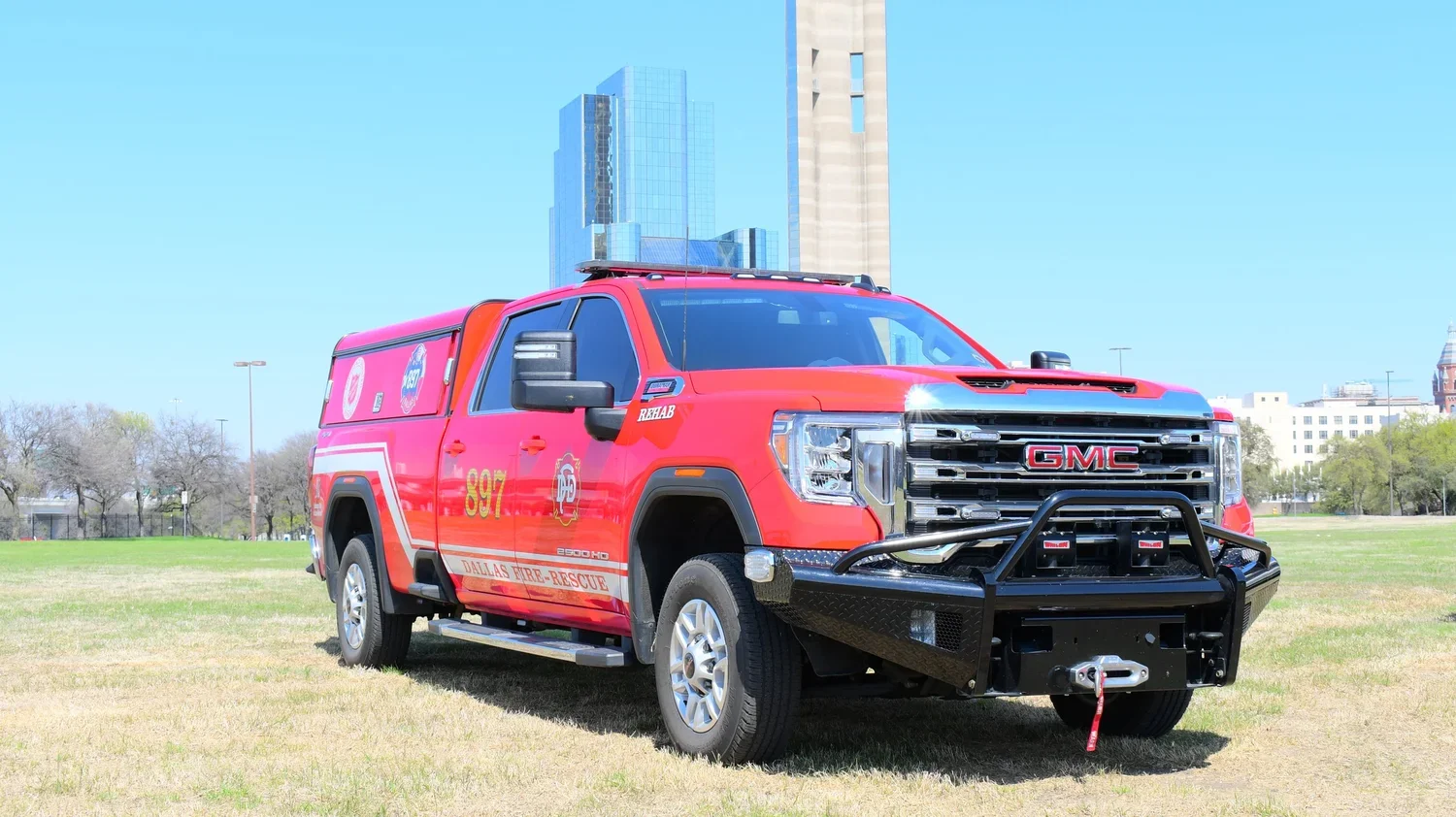 Red fire rescue truck parked on grass with city buildings in the background.