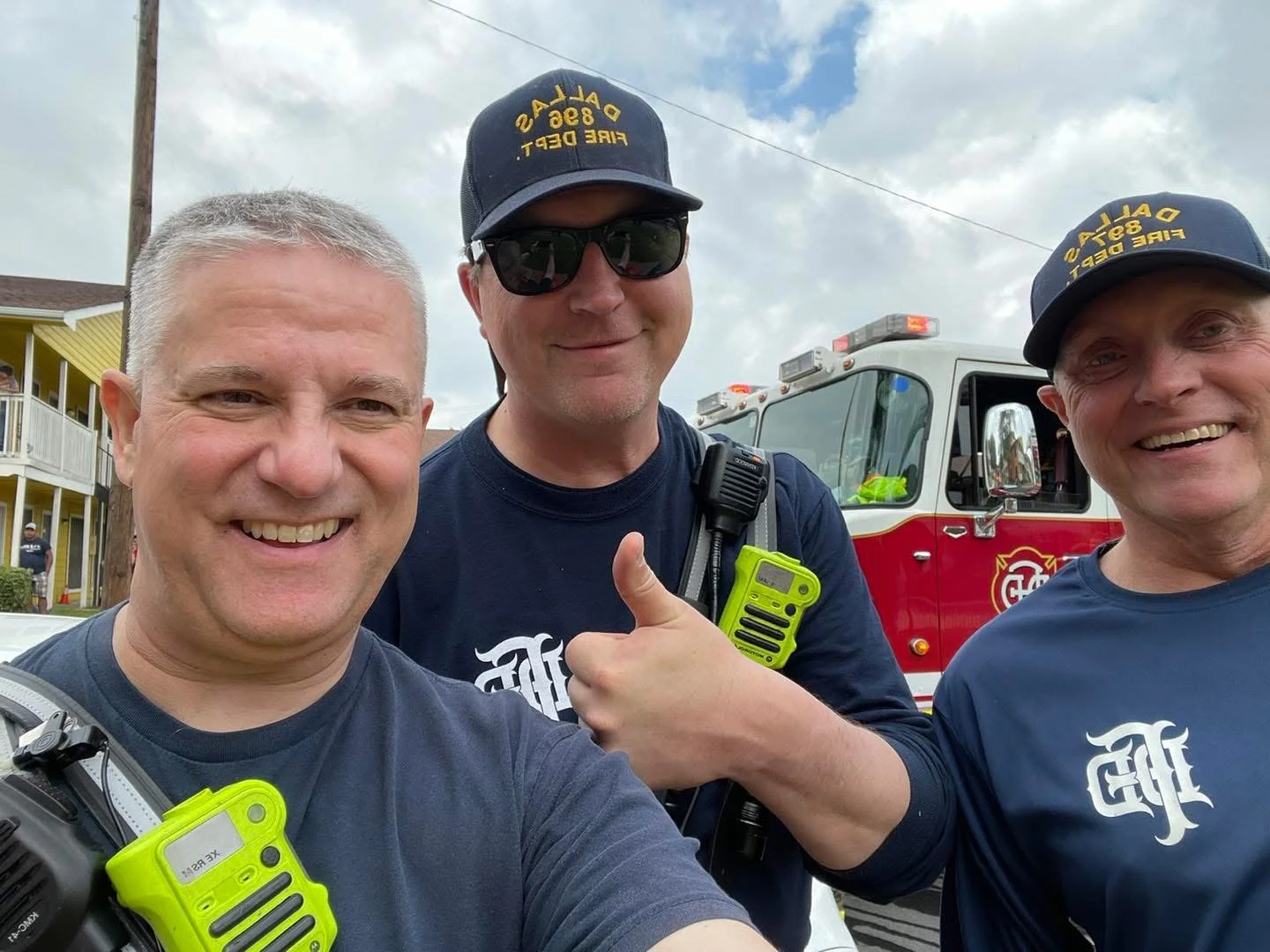 Three smiling men posing for a selfie outdoors in front of a fire truck, dressed in fire response gear with radios, one giving a thumbs-up, all wearing dark shirts and hats.