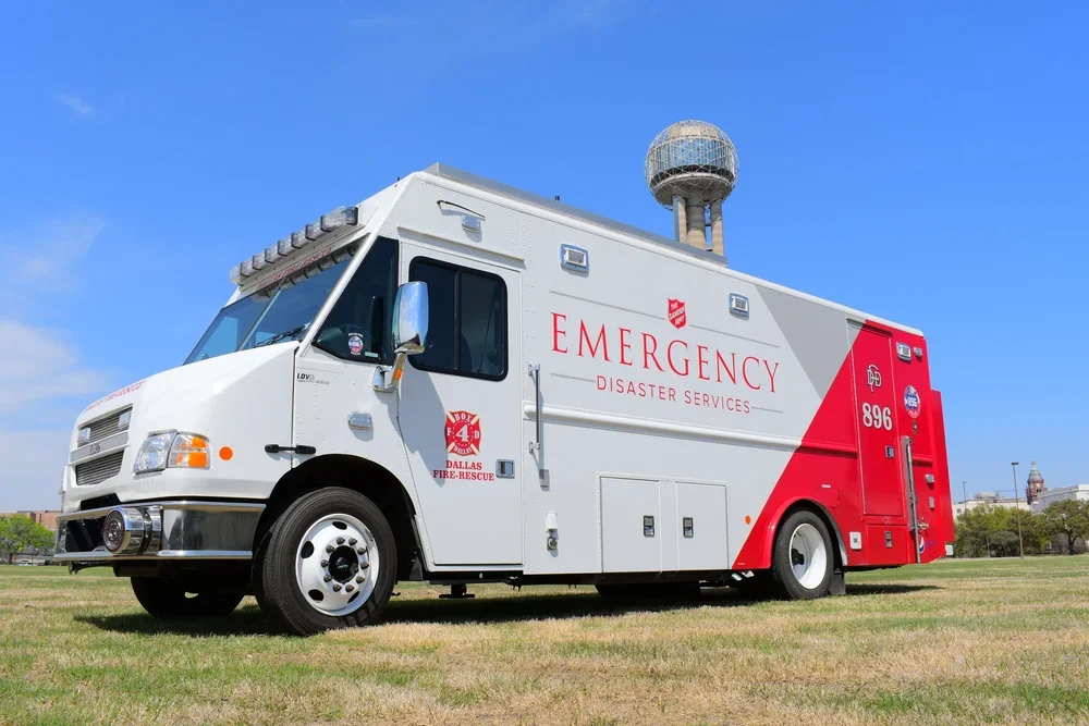 A white emergency disaster services vehicle with a red section, parked on a grassy field, with a small dome-shaped structure visible in the background.