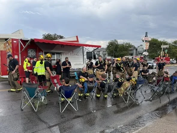 Firefighters and emergency personnel sit and stand around a food truck in a parking lot under a cloudy sky, some wearing helmets and safety vests, with cars and buildings in the background.