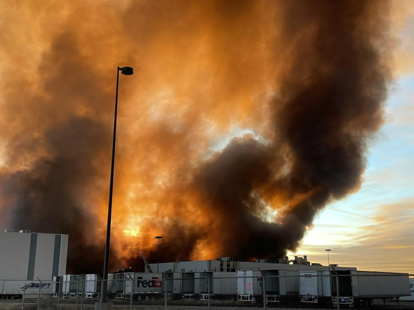 Large black and orange smoke billowing from a building or structure, with several trucks and a chain-link fence in the foreground, and a sunset in the background.
