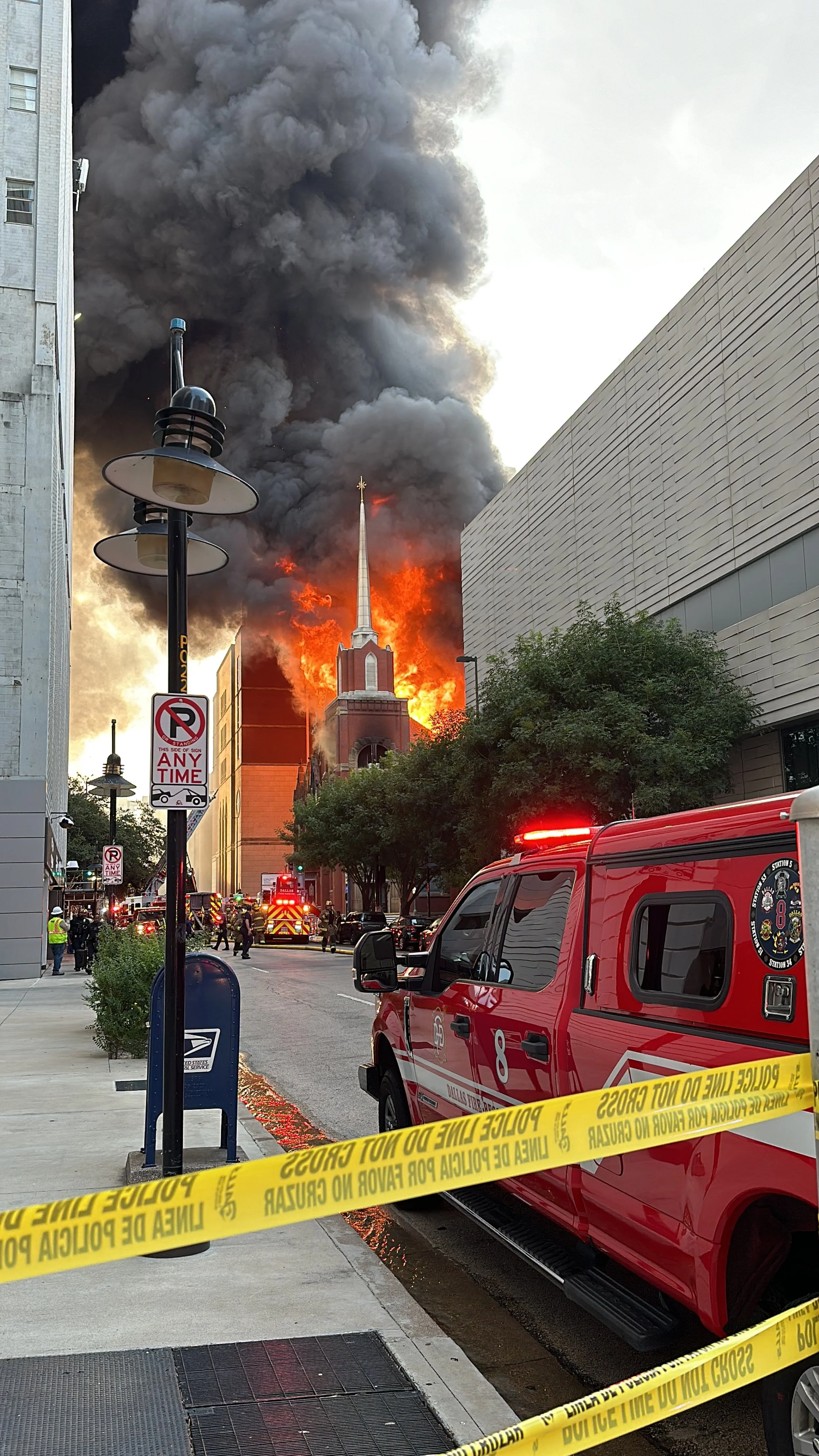 A church on fire with large flames and thick black smoke billowing into the sky, while emergency vehicles and personnel respond on the street in front of the building.