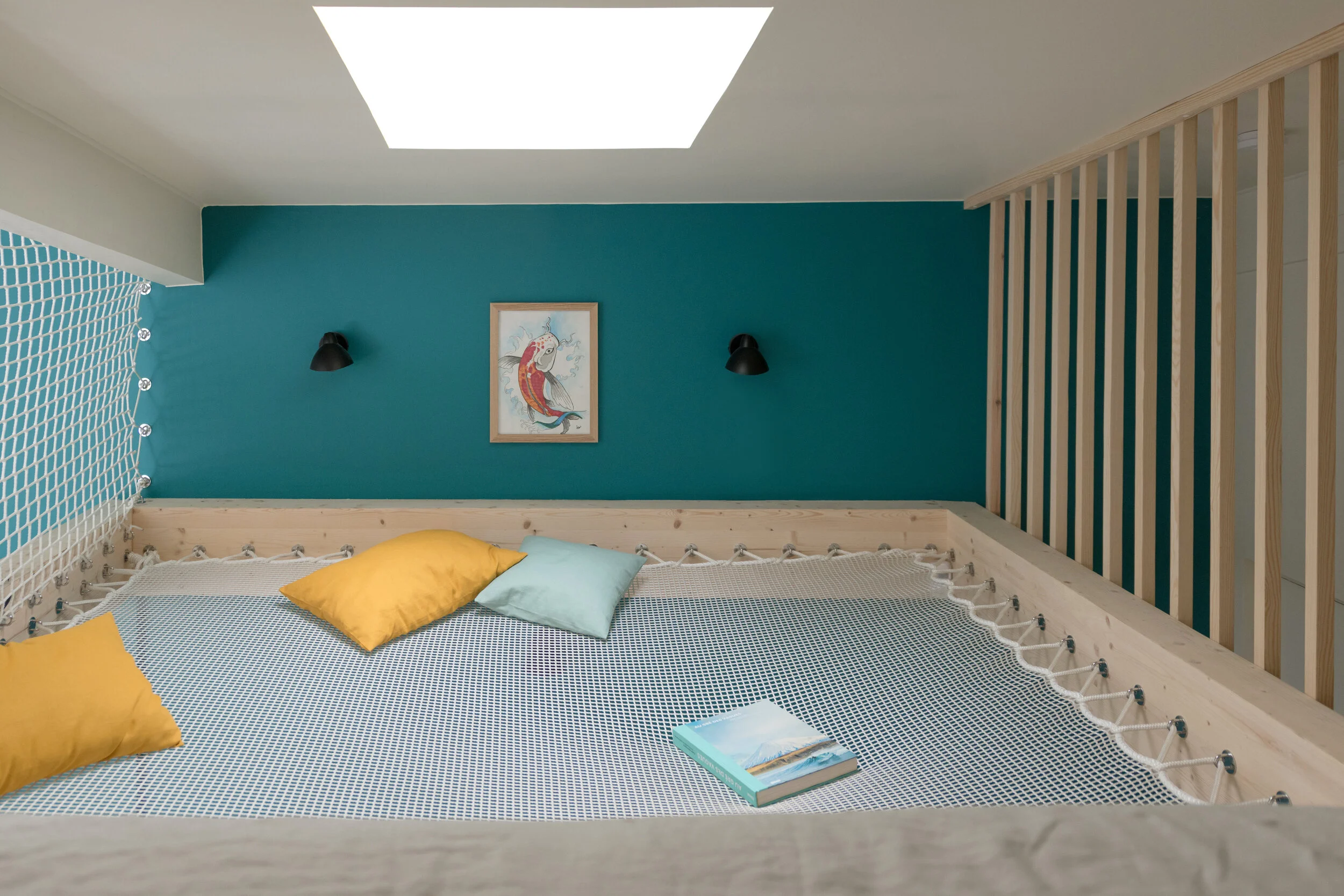 Un espace de détente avec un filet suspendu sécurisé par des cordes, deux coussins jaunes et un coussin bleu clair. Un livre repose sur le filet. Un mur bleu turquoise est décoré d'une illustration encadrée de poissons. Deux appliques noires sont fix