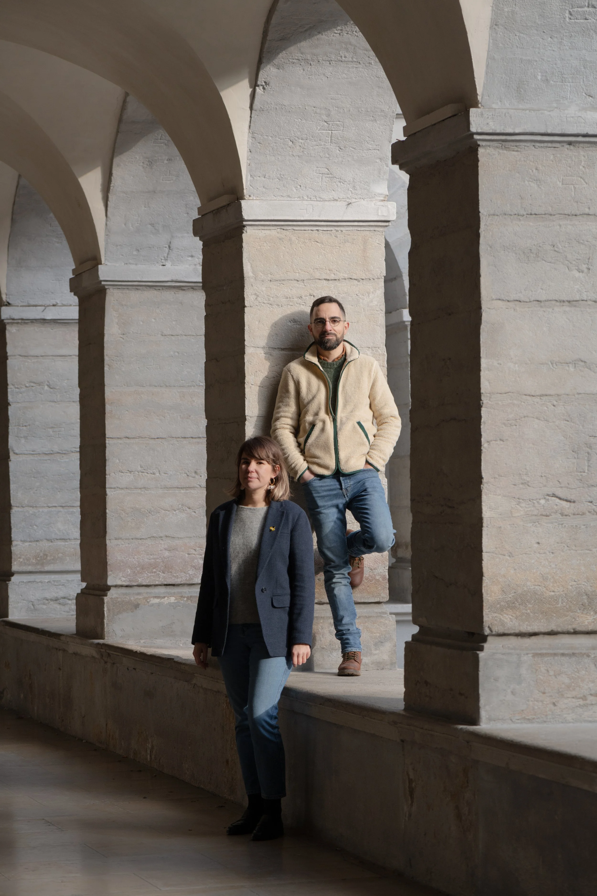 Deux personnes posent sous une arcade en pierre. L'une est debout, l'autre est appuyée contre une colonne.
