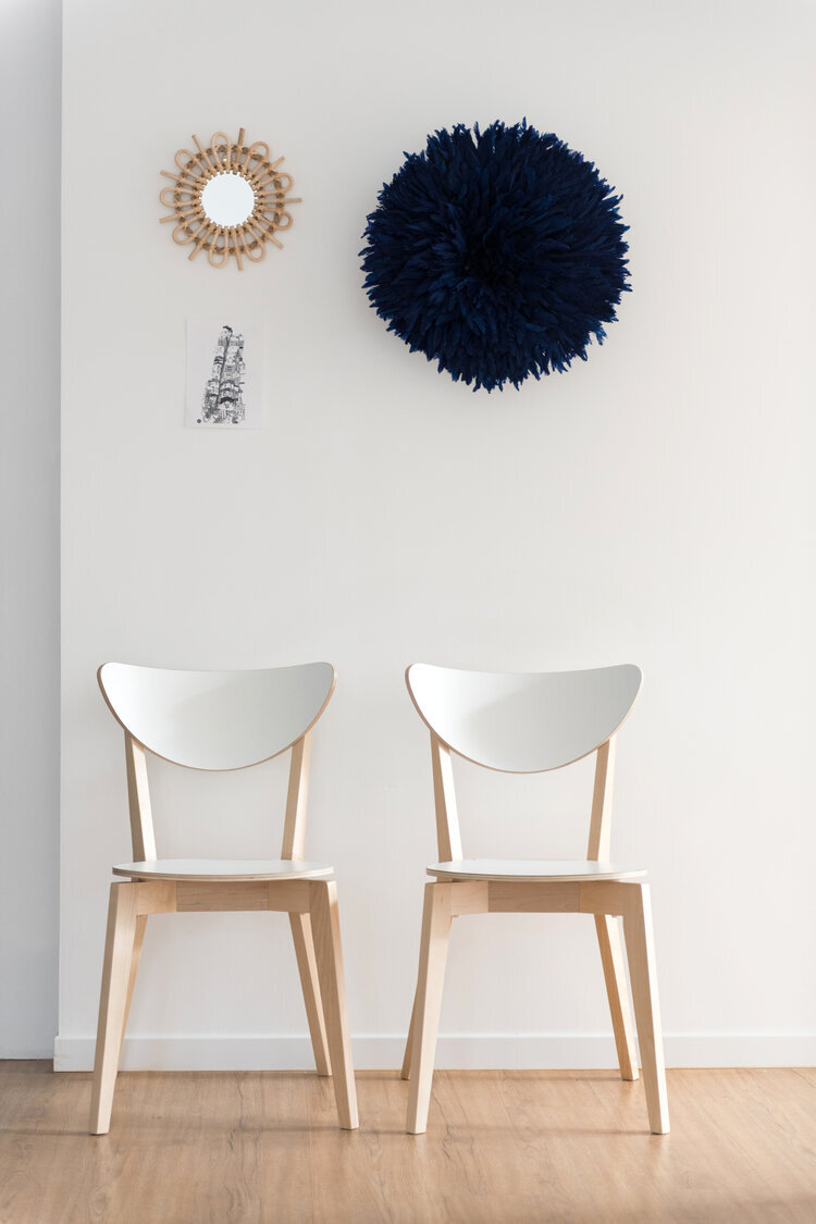 Deux chaises en bois clair devant un mur blanc avec un miroir rond à cadre en osier, un grand ornement mural bleu foncé, et une petite photo en noir et blanc.
