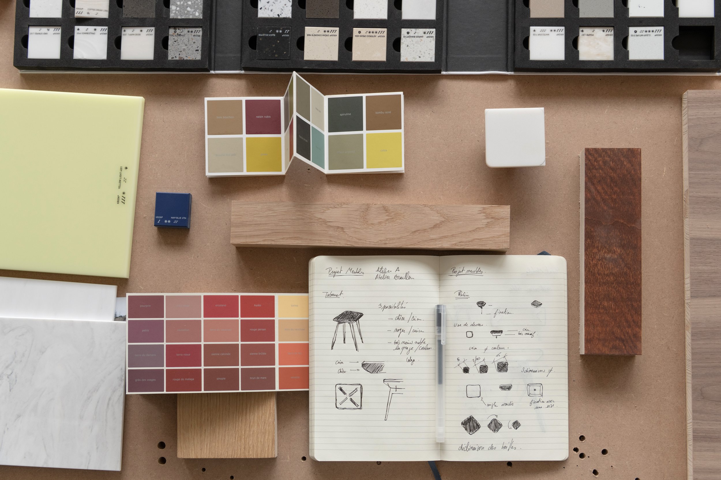 Échantillons de matériaux, nuanciers de couleurs, carnet de croquis, bois, et cube blanc sur une table.