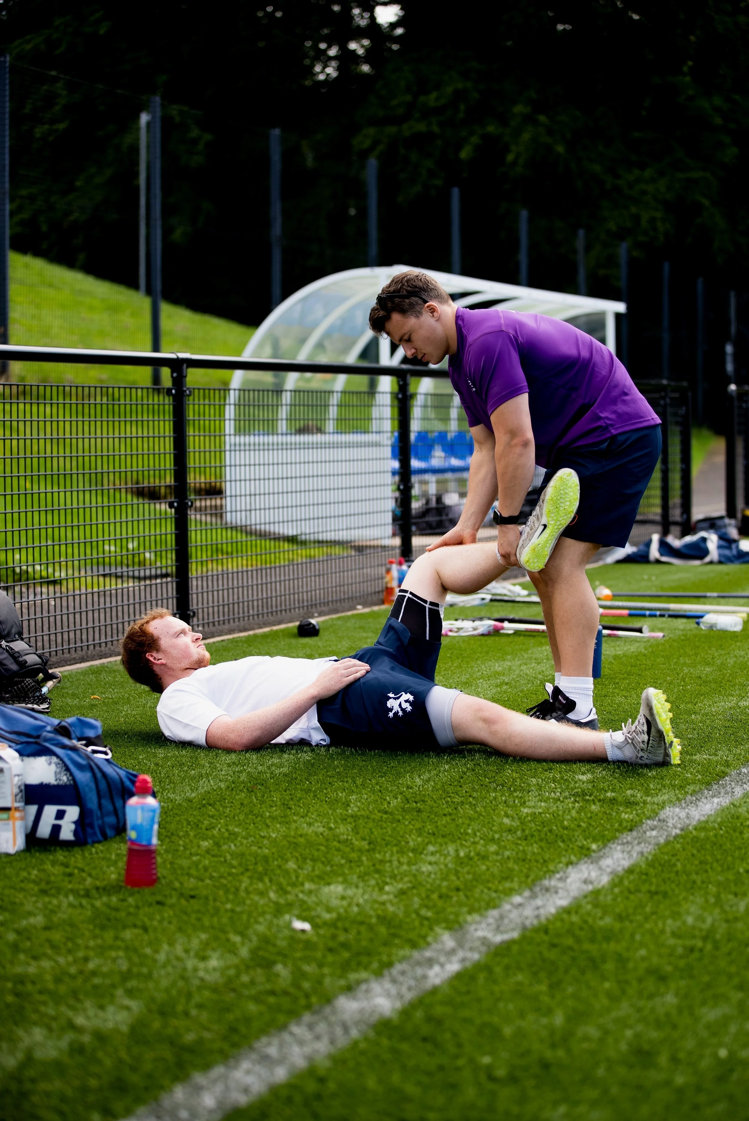 A male sports trainer in a purple shirt and shorts helping a male athlete lying on the grass field, with sports equipment around, during daytime.