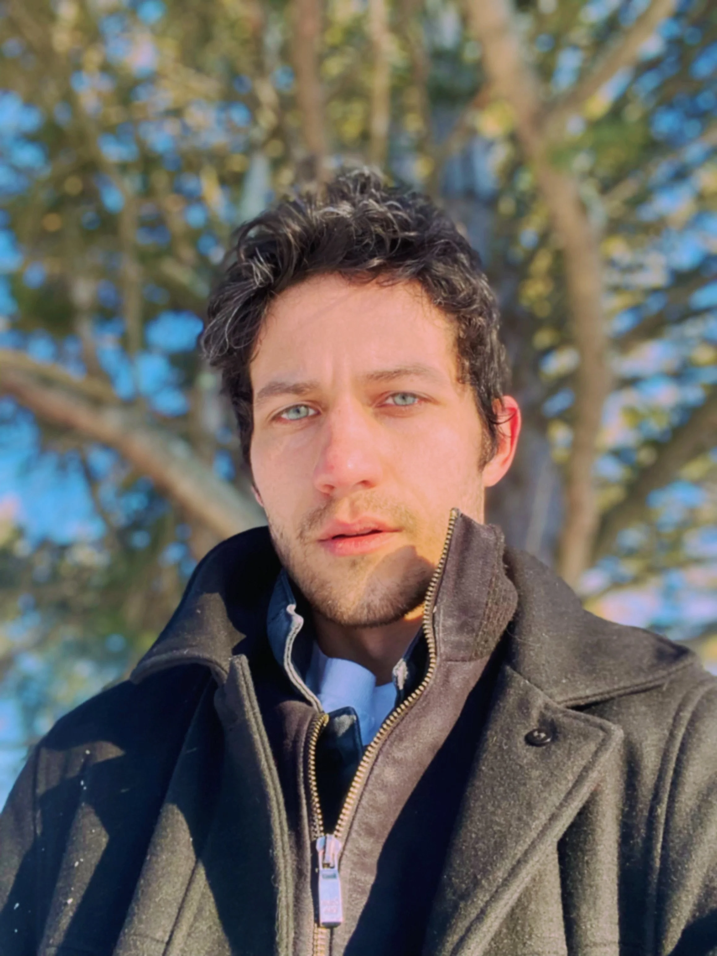 A young man with curly dark hair and blue eyes taking a selfie outdoors in front of a large tree with green leaves and a blue sky background.