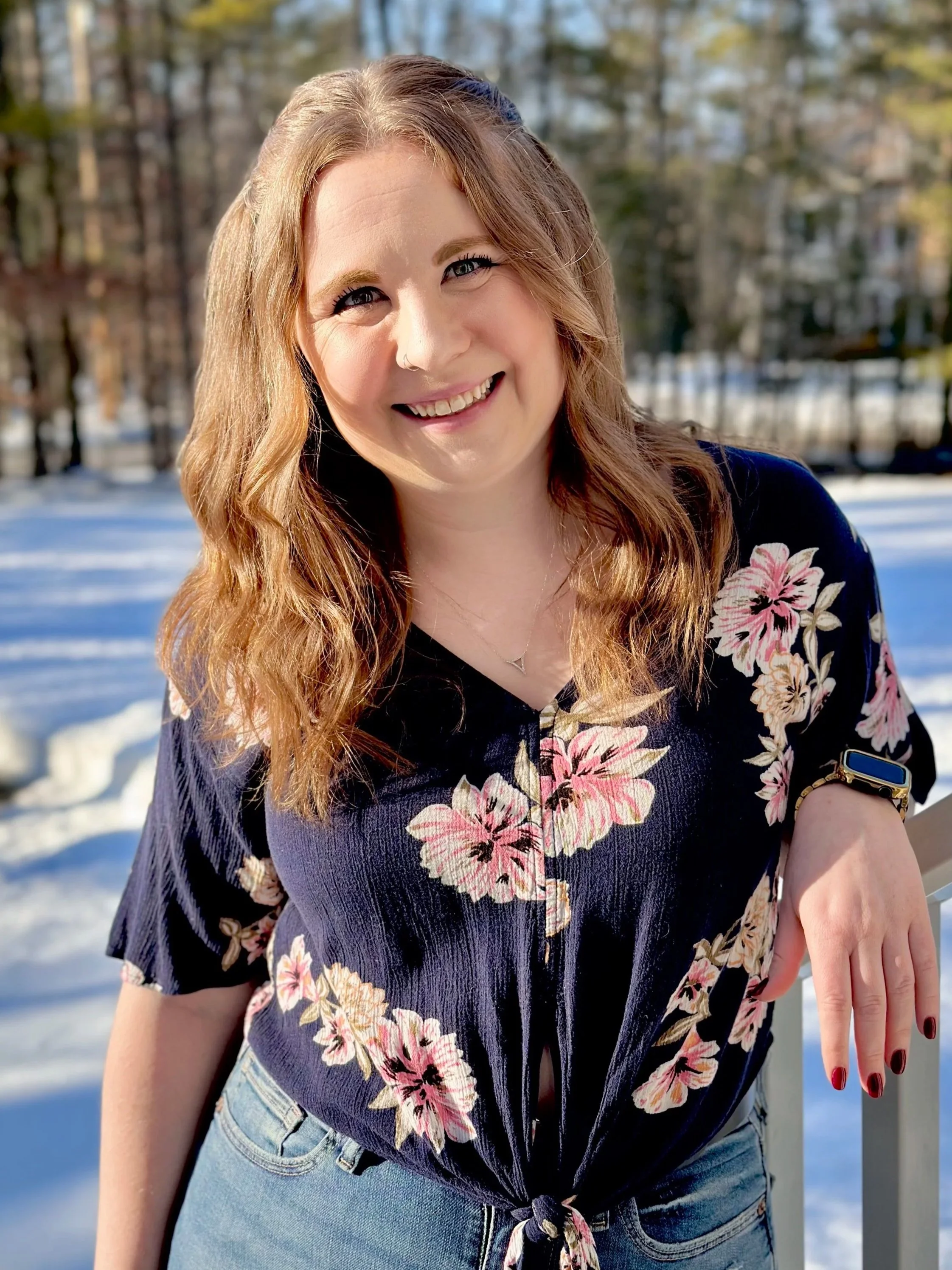 A young woman with wavy red hair smiling outdoors in winter, wearing a navy floral top, light blue jeans, and a smartwatch, standing on a snowy landscape with trees in the background.
