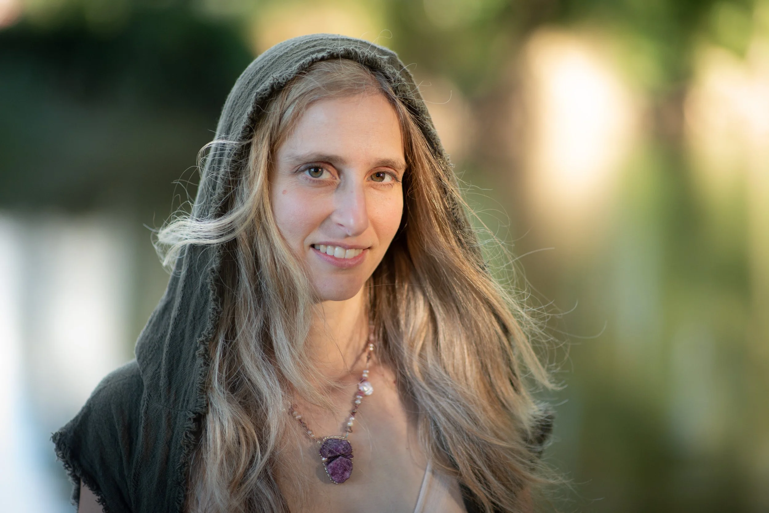 A woman with long blonde hair wearing a necklace with purple stones and a gray hooded shawl, outdoors with blurred green and yellow background.