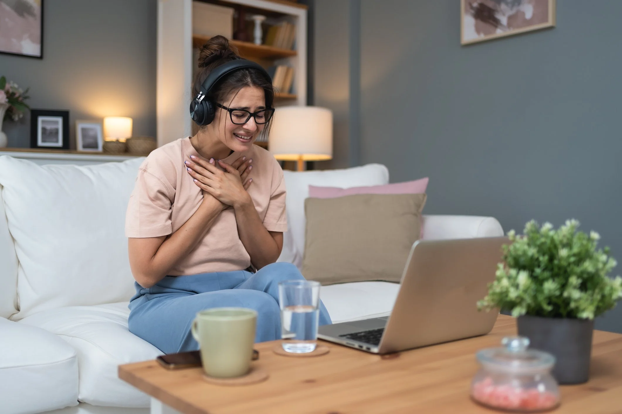 Woman with glasses and headphones clutching her chest while seated on a white couch in a living room, with a laptop, a glass of water, a mug, and various decor items on a wooden table.