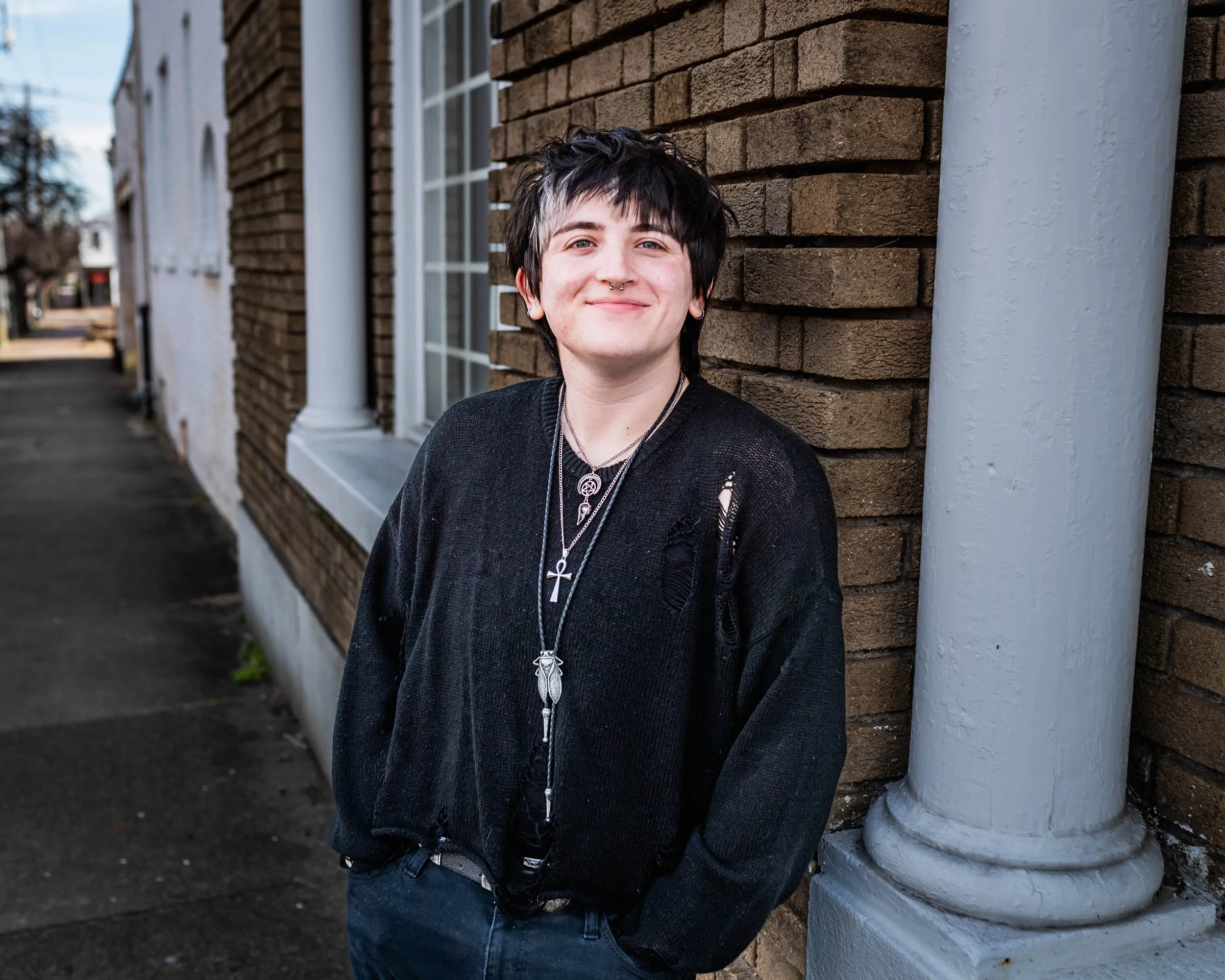 Young person with short dark hair and piercings, smiling, standing outside near a brick building with large windows and a white pillar.
