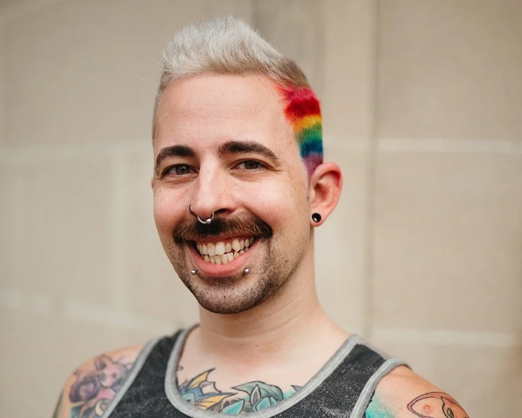 A smiling man with tattoos, rainbow-colored hair on the side of his head, multiple facial piercings, and a tank top, standing against a plain background.