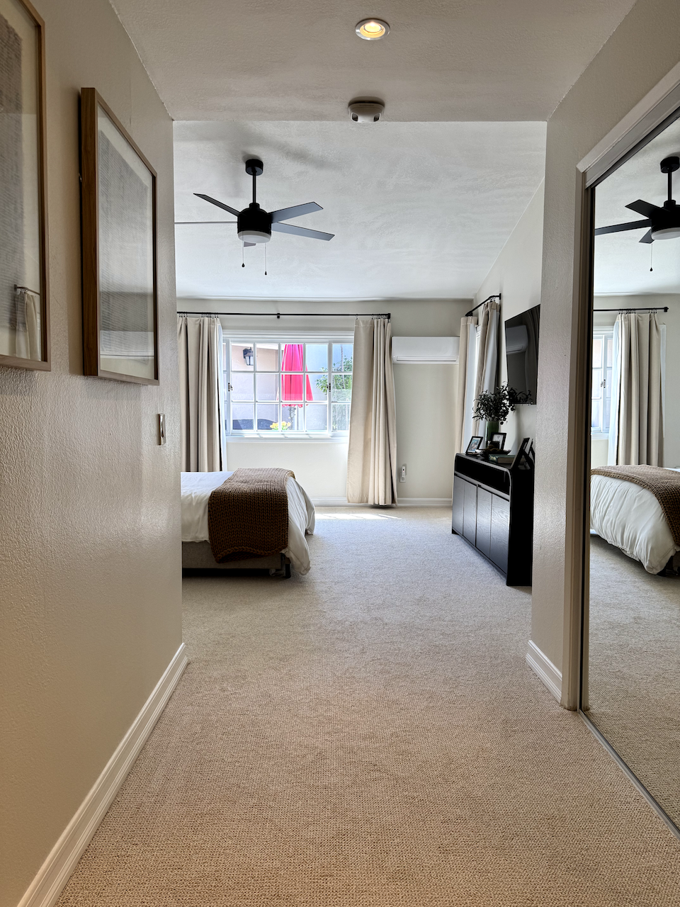 A hotel bedroom with a bed, window with curtains, ceiling fan, wall-mounted air conditioner, TV on a console, and a mirrored closet door.