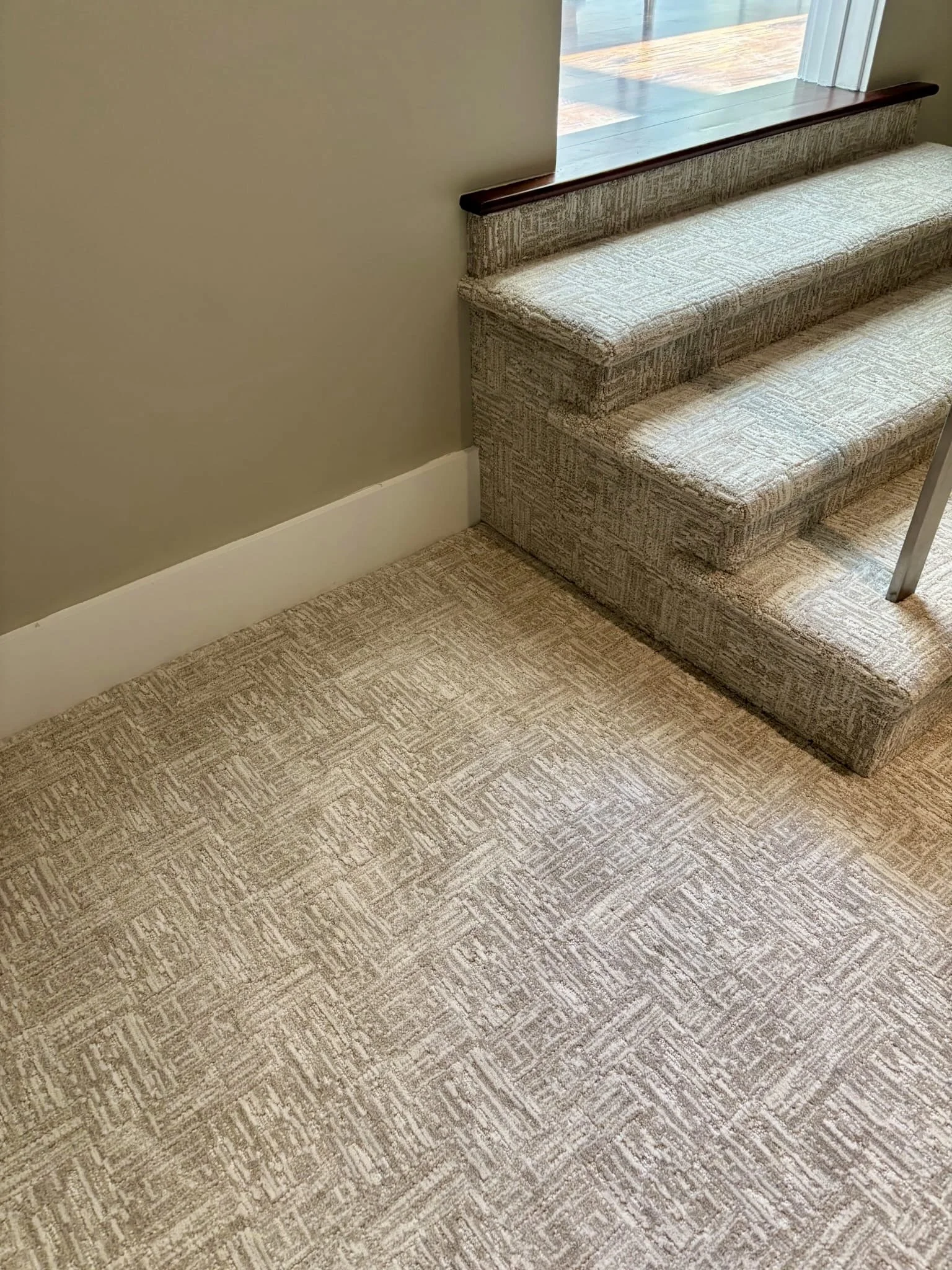 Interior of a room showing a carpeted staircase with beige patterned carpet, a beige wall, and a window with natural light.