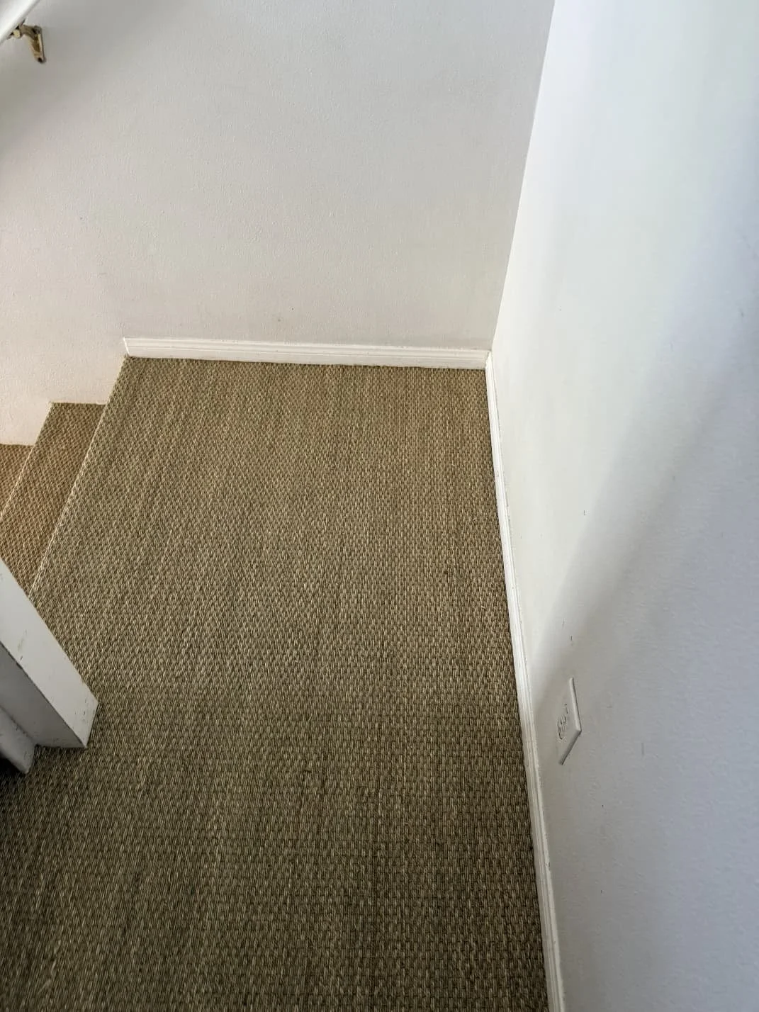 Photo of a corner in a room showing a beige carpeted floor, white walls, a staircase, and an electrical outlet.