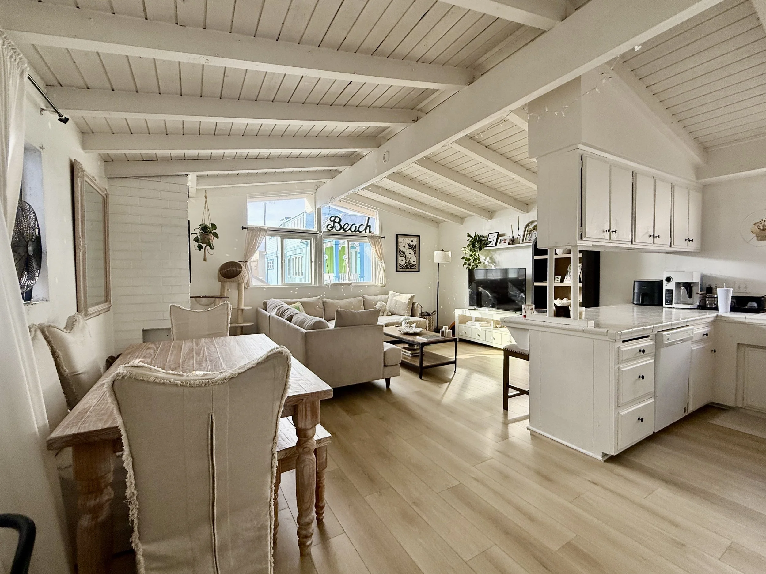 Open-concept living room and kitchen with white walls, wood flooring, and exposed wood ceiling beams. The living room has a beige sectional sofa, a coffee table, and a TV stand with a flat-screen TV. The kitchen has white cabinets, a countertop with 