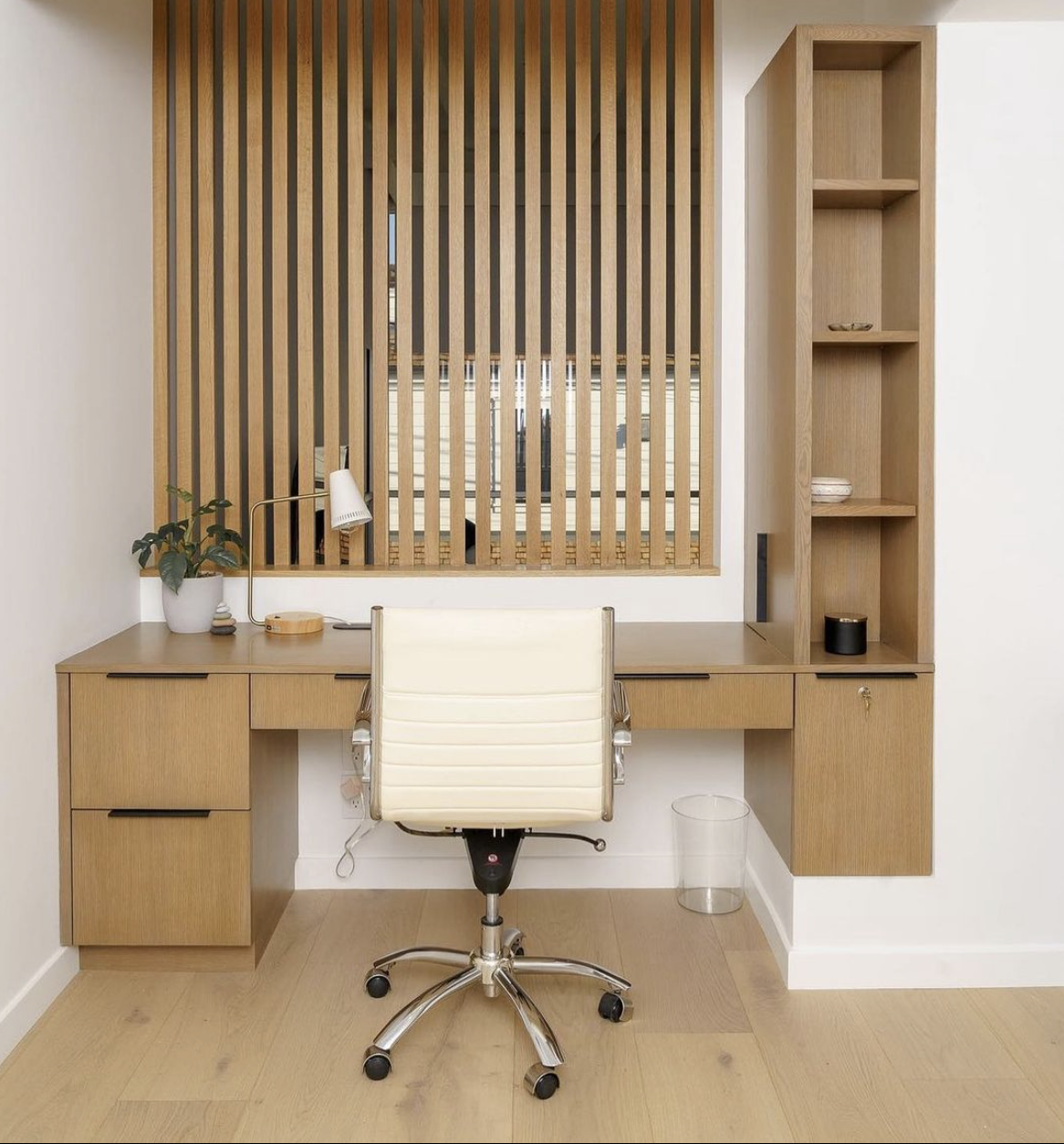 Minimalist home office with a wooden desk, white office chair, potted plant, and wooden slat partition with shelving on the right and window behind.
