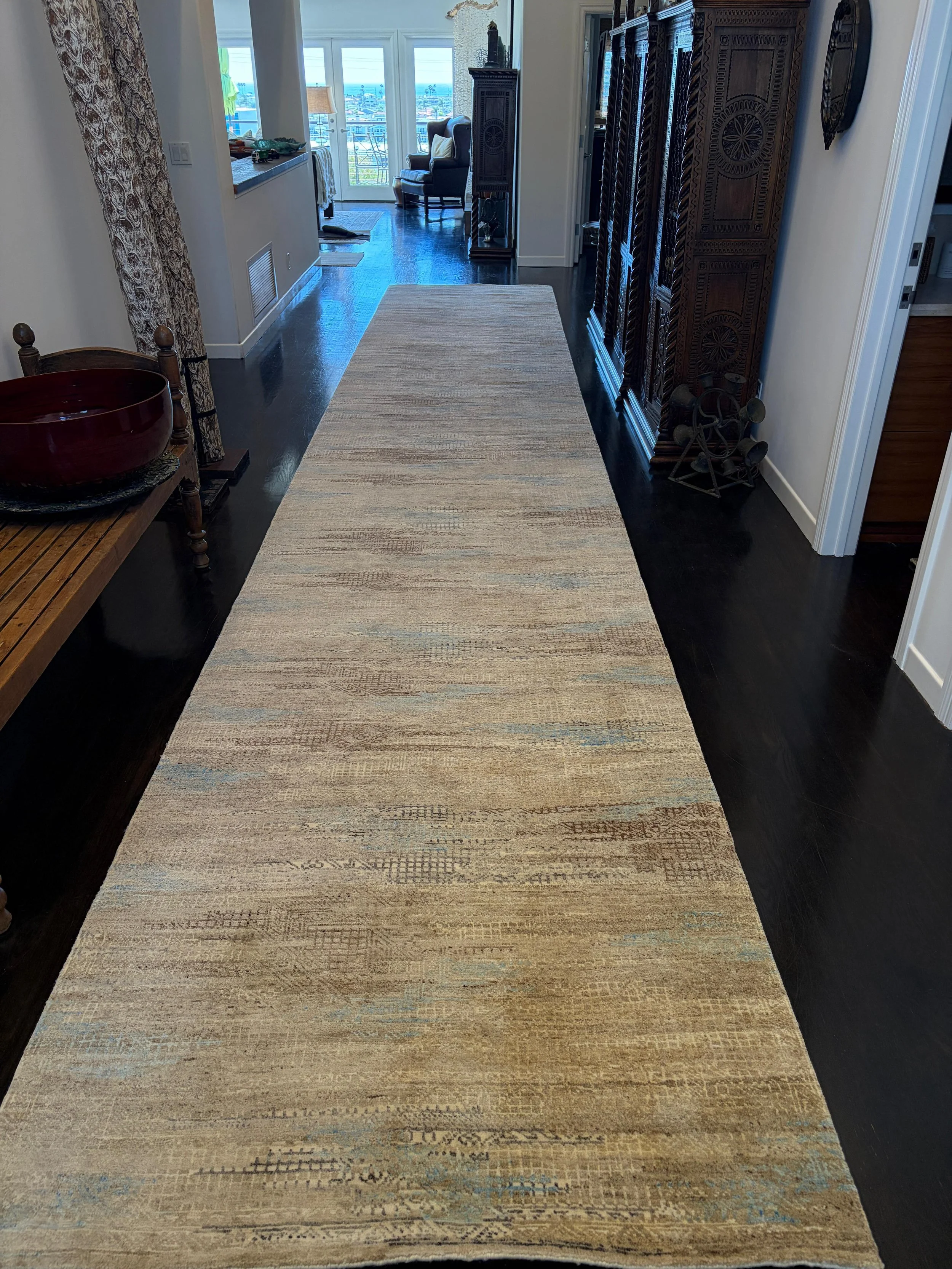 A hallway with a beige and gray rug on dark hardwood floor, leading to a bright room with glass doors and ocean view, with furniture and decorative items.