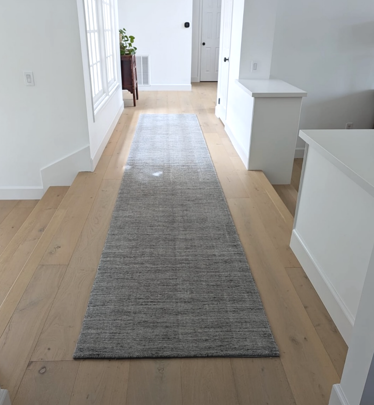A long gray runner rug on light wooden flooring in a bright hallway with white walls, and a small table with a plant in the background.