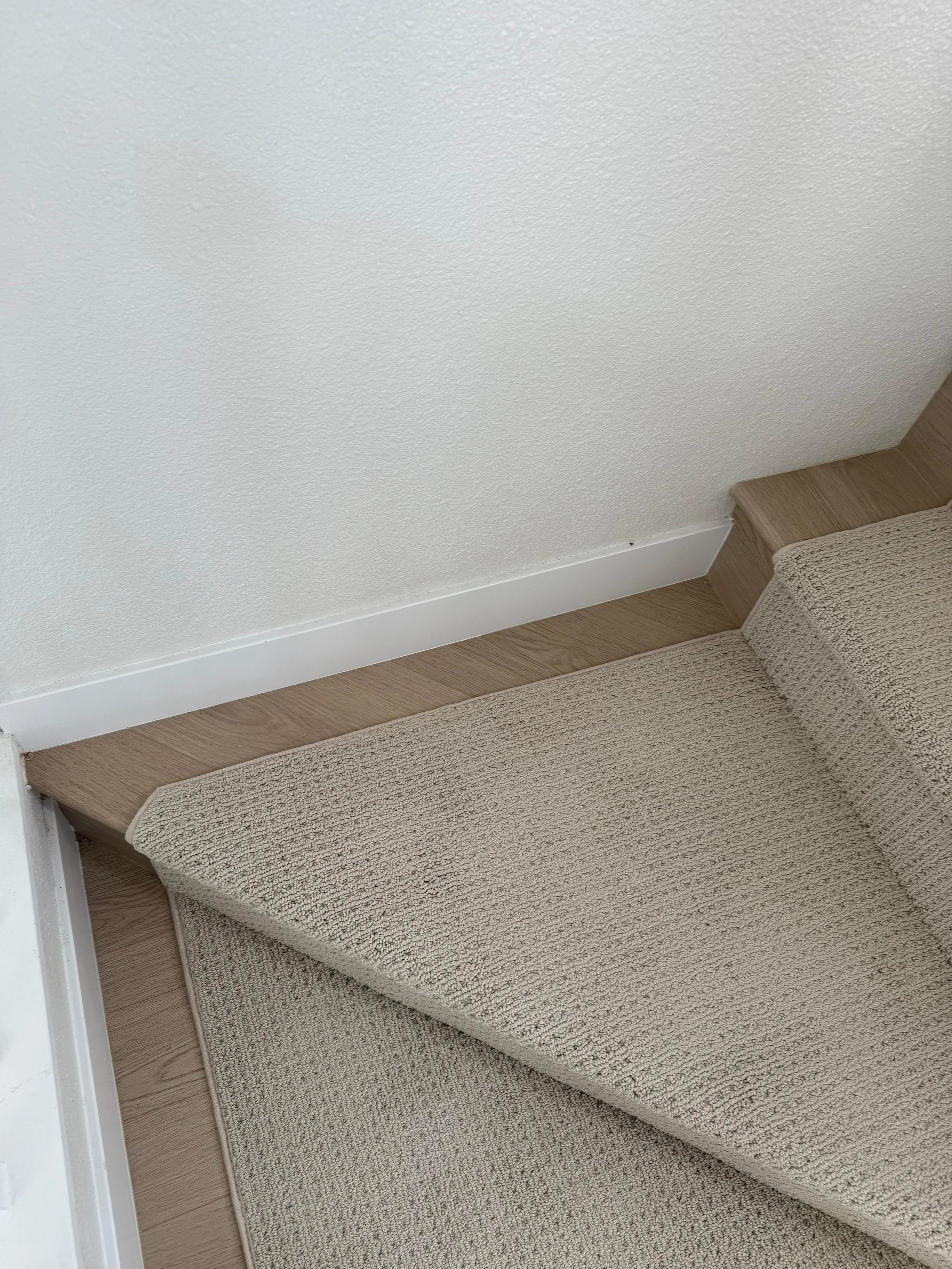 Close-up of a beige carpeted staircase landing with a carpeted step and a section of hardwood floor, near a white wall with a baseboard