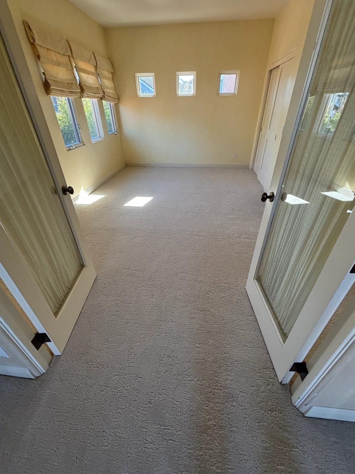 Empty room with beige carpet, yellow walls, small rectangular windows, and closed closet doors, seen through glass double doors.