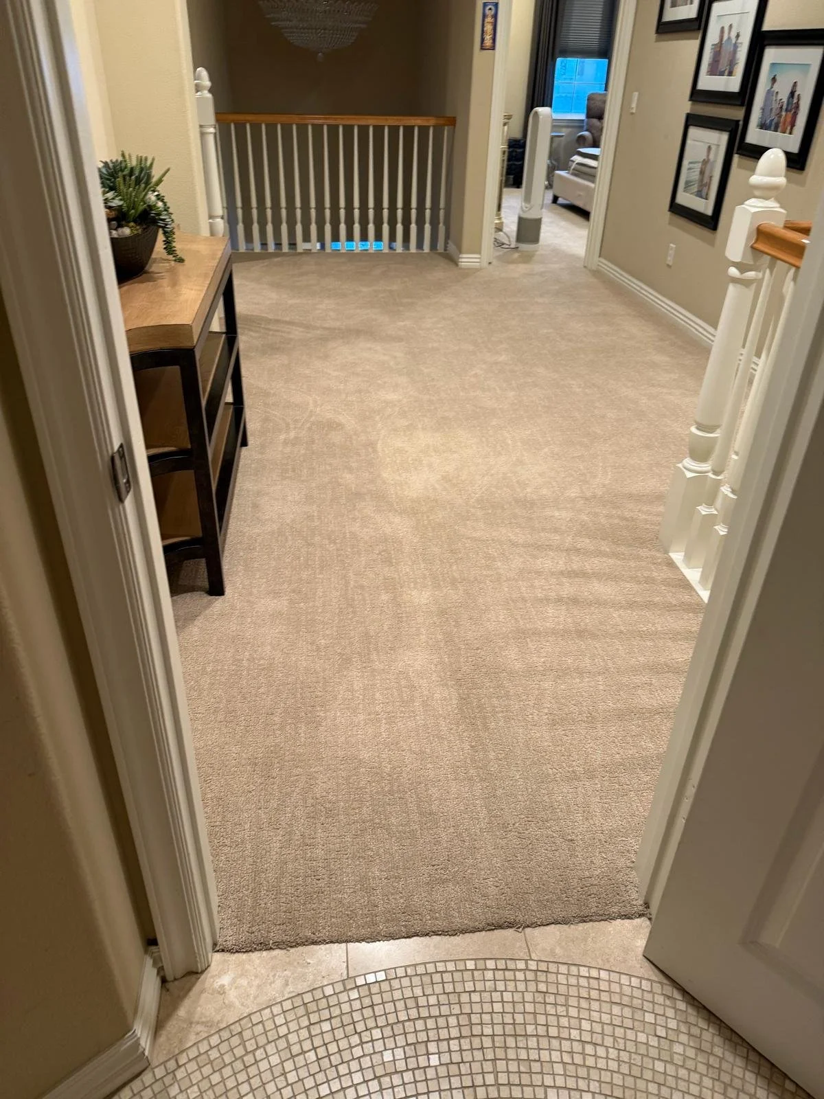 Interior view of a hallway in a house, showing a doorway with beige carpet, a small black shelf with a green plant on top, framed pictures on the wall, and a staircase railing leading to an upper level.