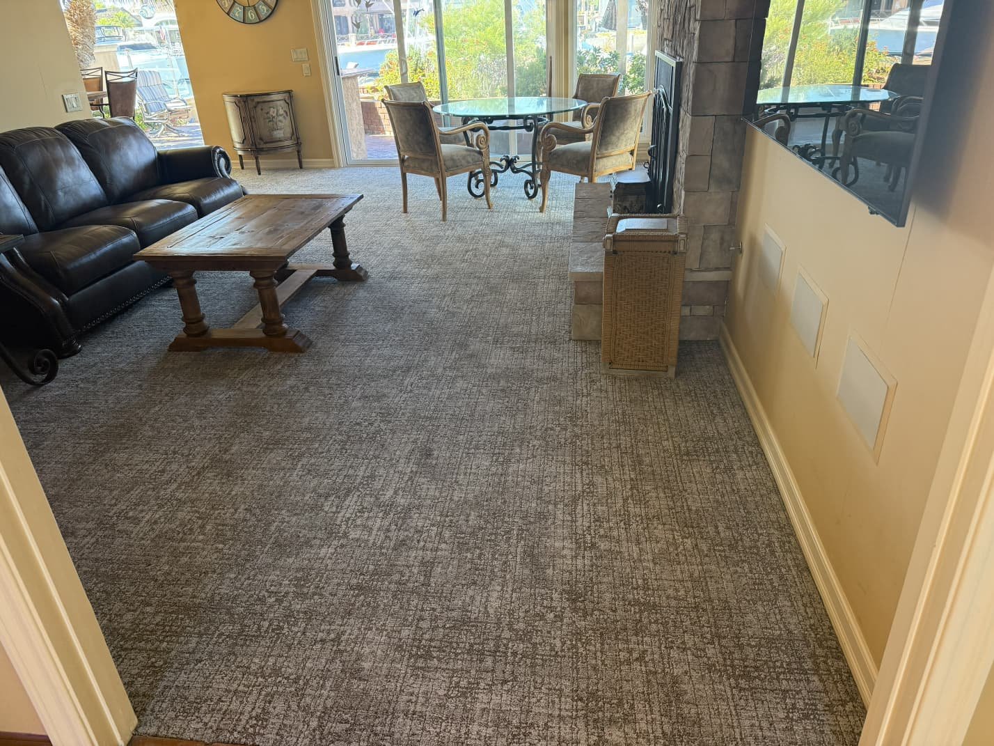 Living room with dark leather sofa, wooden coffee table, glass dining table with four chairs, and a large flat-screen TV mounted on the wall. Large windows and sliding glass door lead to an outdoor patio.