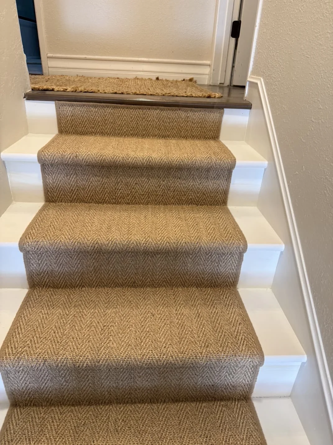 Staircase with four beige carpeted steps, white risers, wood trim, and a brown doormat at the top