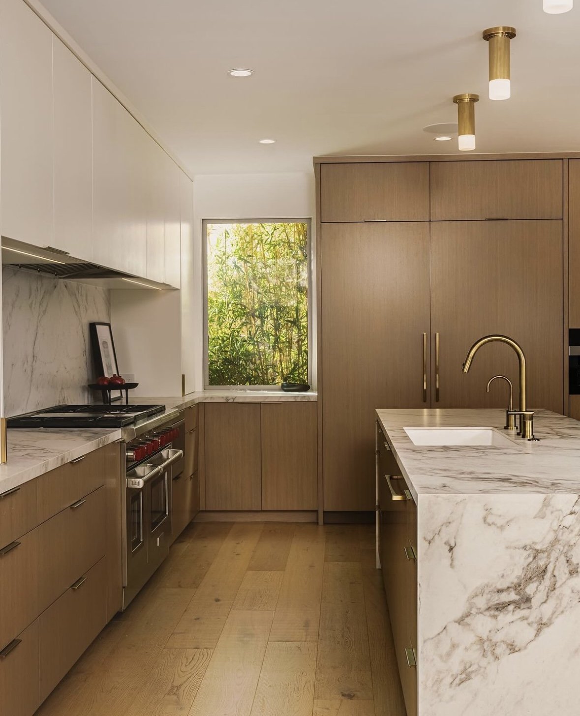 Modern kitchen with wooden cabinets, a marble countertop island with a sink, a window with greenery outside, and ceiling lights.